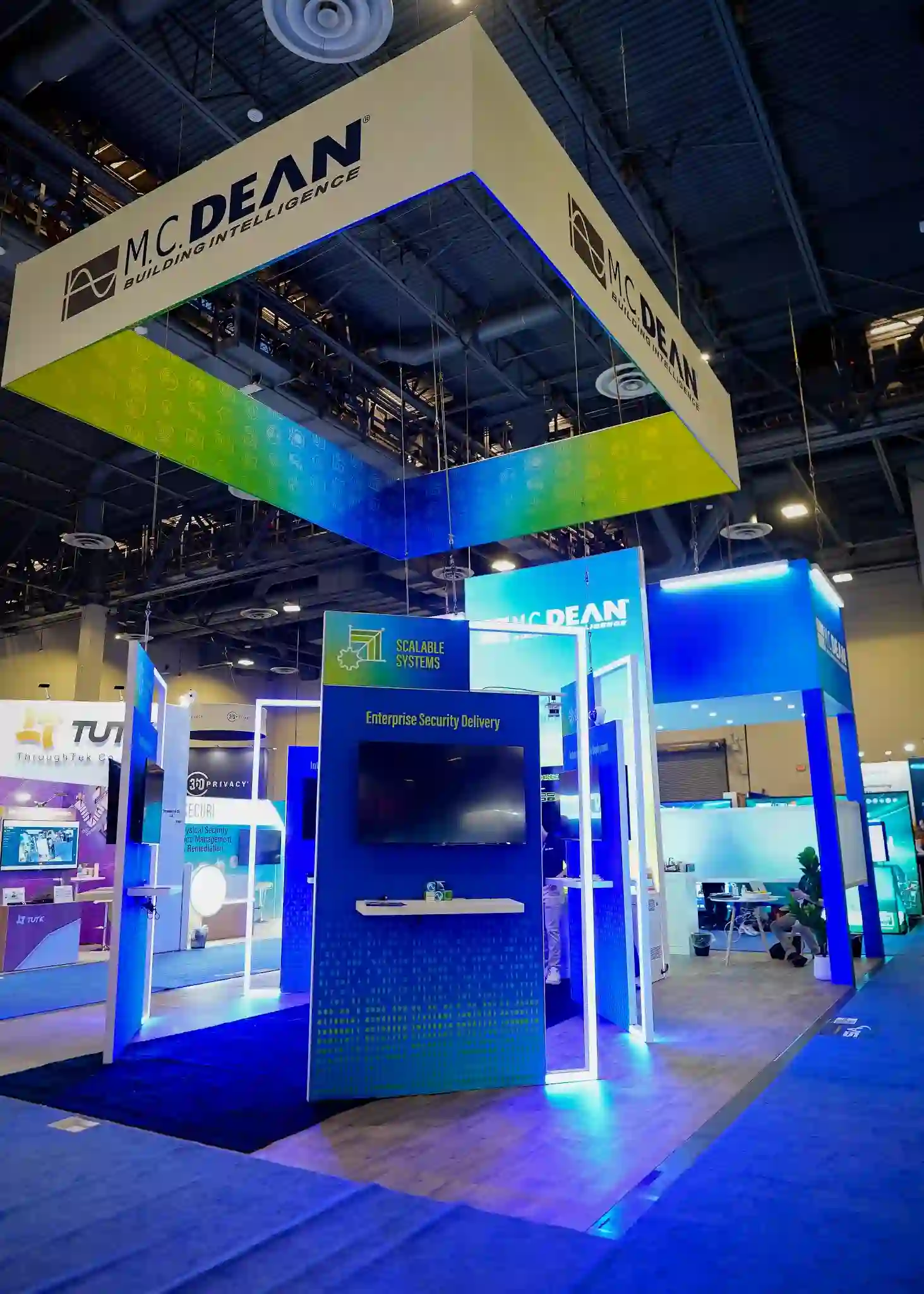 Angled upward view showing scale of hanging fabric sign with blue-to-green gradient, Scalable Systems kiosk with globe network graphic, and Building Intelligence branding against dark hall ceiling