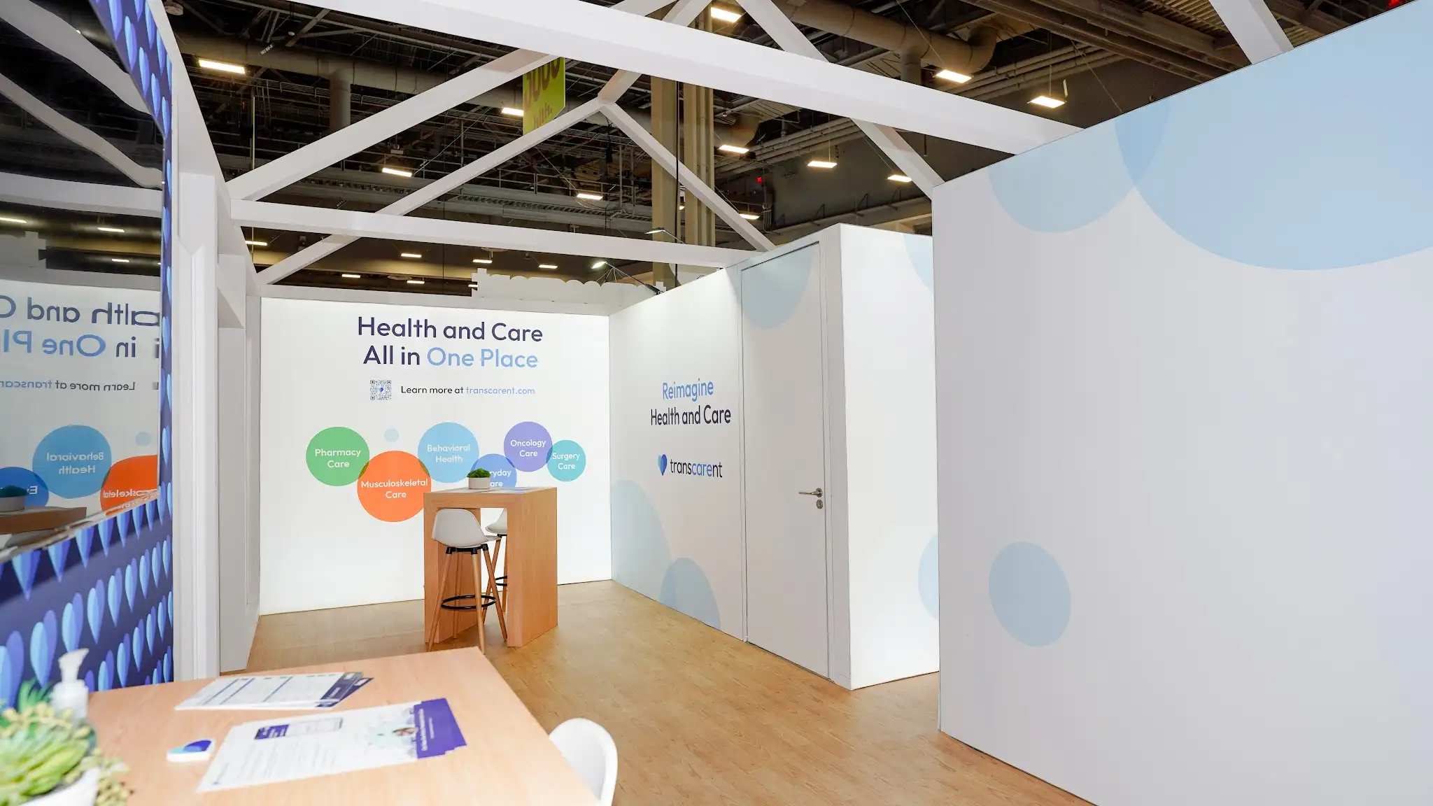 Close-up interior view looking into the front opening of the Transcarent house booth at HLTH 2023 showing left interior front panel with Health and Care All in One Place headline in dark navy and One Place in larger Transcarent brand blue, QR code with Learn more at transcarent.com, multicolored bubble chart infographic with six overlapping circles Pharmacy Care in green Musculoskeletal Care in orange-red Behavioral Health in purple Oncology Care in teal Surgery Care in blue Everyday Care in blue, back white glowing panel with TV monitor showing purple-blue content, deep blue indigo interior accent wall with lighter blue repeating heart pattern on right, modern white barstool with wooden legs and small green succulent plant on wooden table, white structural columns flanking view, warm wood flooring