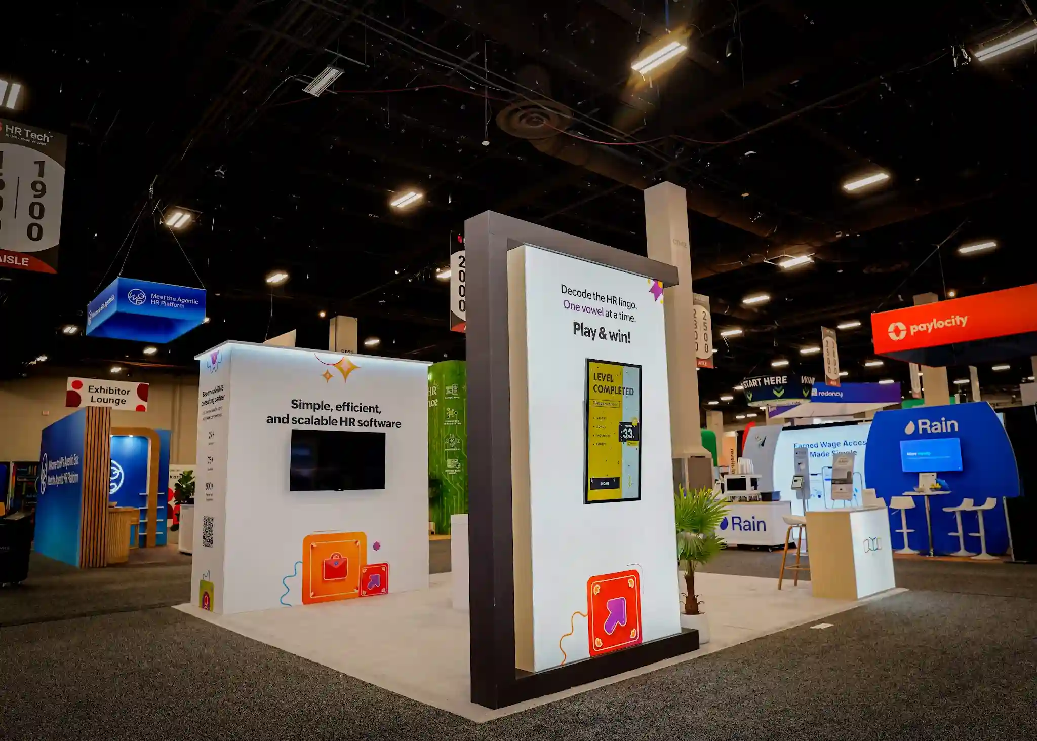 Close-up of the Zoho interactive gaming tower showing Decode the HR Lingo One Vowel at a Time Play and Win header, large vertical touchscreen with vivid yellow Level Completed game screen and score of 33, dark charcoal frame structure, orange briefcase icon at the base, adjacent product messaging panel with Simple efficient and scalable HR software headline and flat-screen TV, wooden bar stool, and neighboring booth signage