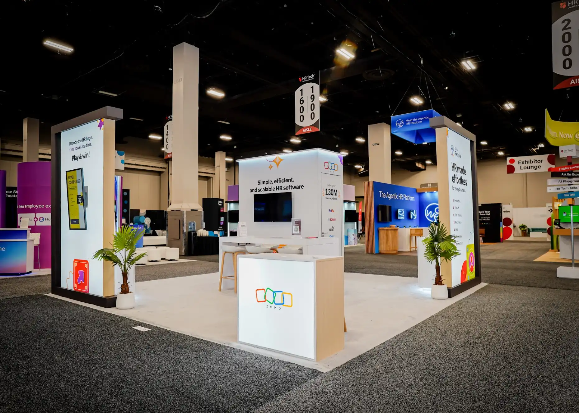 Wide-angle overview of the Zoho 20 by 20 island booth at HR Tech 2025 showing white architectural gateway columns with dark charcoal framing, interactive gaming display tower with yellow Level Completed screen, backlit Zoho logo reception counter, tropical palm plants in white planters, orange bar stools at white bar-height tables, and bright white custom flooring contrasting with the dark convention center carpet