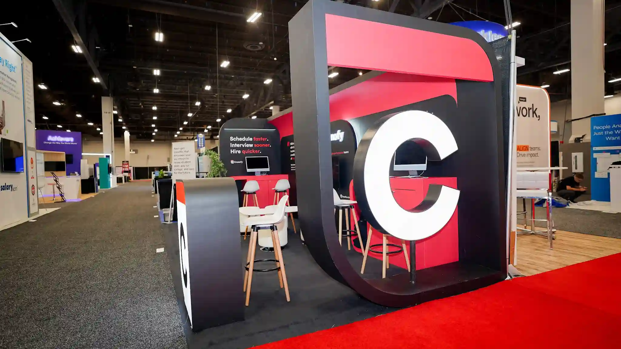 Side-angled view of the Cronofy 10x20 ft island booth at HR Tech 2025 looking across the show floor from the right side showing the full row of booths in context, large vertical C icon tower prop on far right prominent in foreground, booth interior with seating counters and back wall fully visible, dark charcoal carpeting inside booth, red countertops and cabinetry, gloss and matte black structural panel work, clean squared-off edges with curved arch elements, Schedule faster Interview sooner Hire quicker on left panel, Cronofy branding on center panels, neighboring Achievers purple booth salary.com and others visible, broad convention hall ceiling with exposed overhead steel trusses, mostly empty hall indicating pre-show or early setup
