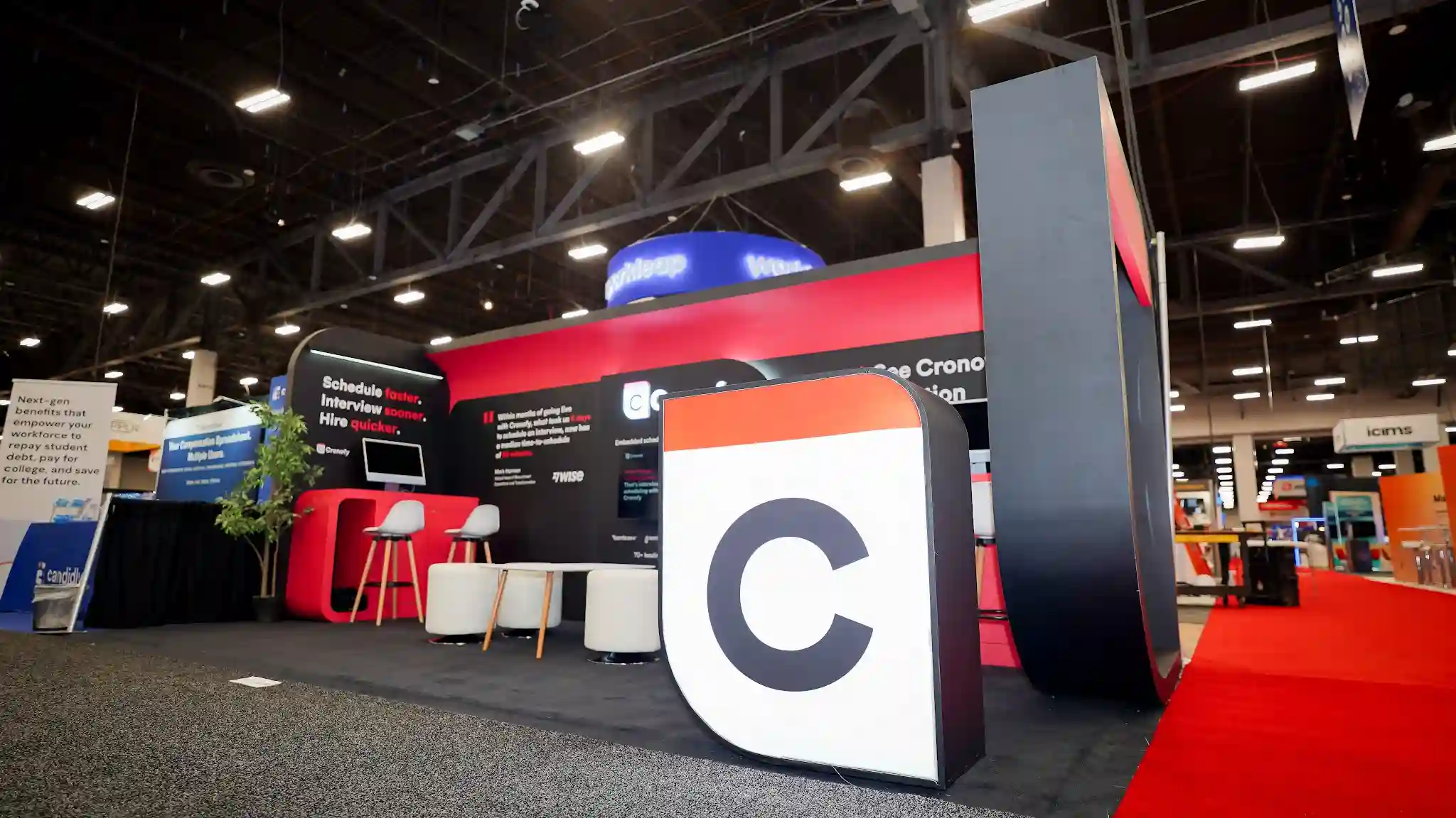 Wide-angle side perspective view of the Cronofy 10x20 ft custom island trade show booth at HR Tech 2025 taken from a lower vantage point to the left-front showing the full island configuration from corner to corner, bold red and black palette with arching pod structures and back wall, large freestanding 3D Cronofy C app icon prop in white with black C letter prominent in lower-right quadrant, Cronofy logo on central back wall, Schedule faster Interview sooner Hire quicker on left panel, See Cronofy in action on right side, neighboring Workleap circular illuminated hanging sign visible, iCIMS booth partially visible to far right, no people present pre-show setup photo, convention center ceiling with industrial lighting