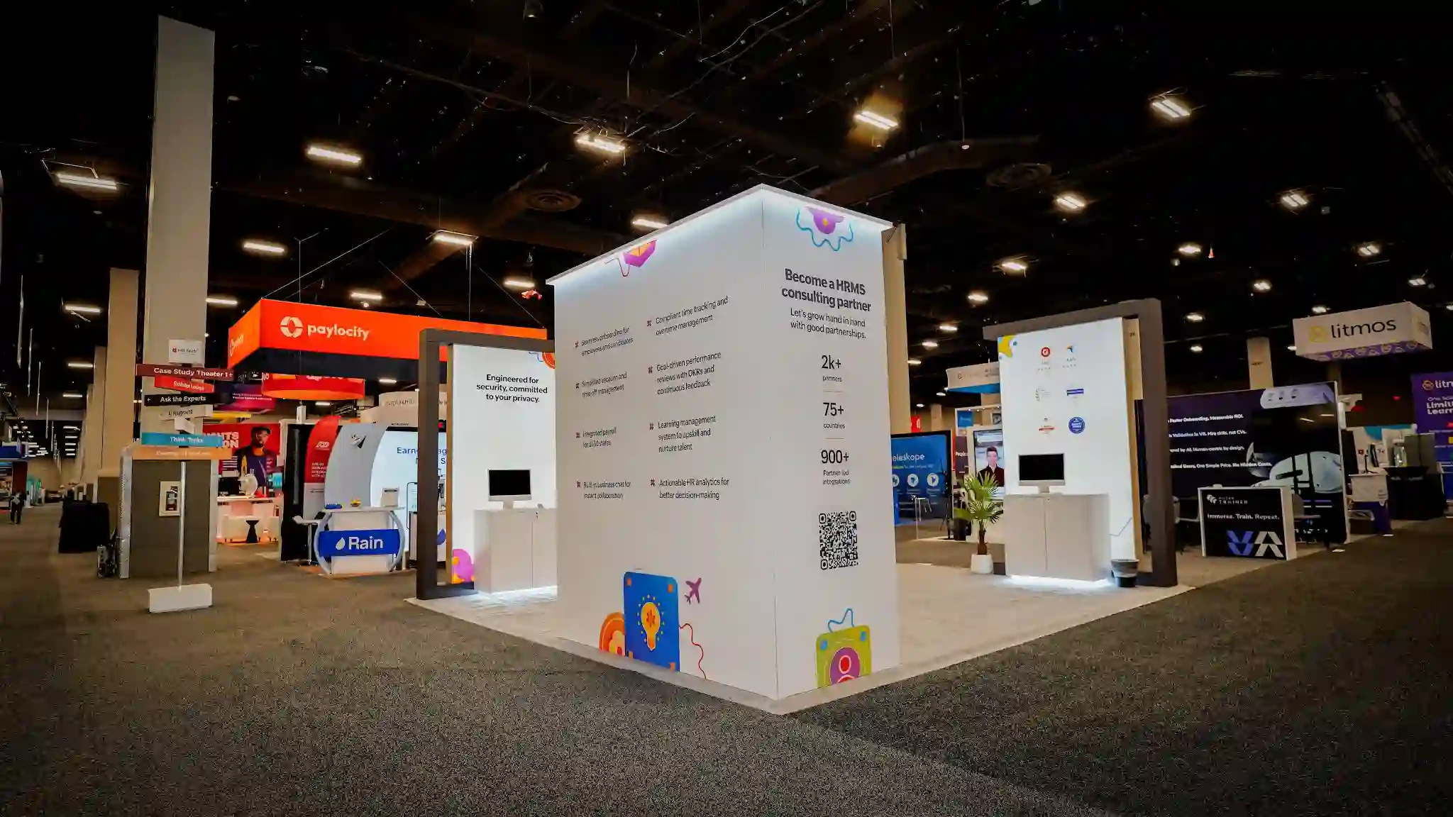 Side-angle view of the Zoho island booth showing the HRMS consulting partner panel with 2k plus partners 75 plus countries 900 plus partner-led integrations stats, Engineered for security committed to your privacy panel, product demo screens mounted on tower columns, colorful illustrated icons including lightbulb teal avatar and shopping bag, white custom flooring, and neighboring Paylocity and Rain booths visible in background