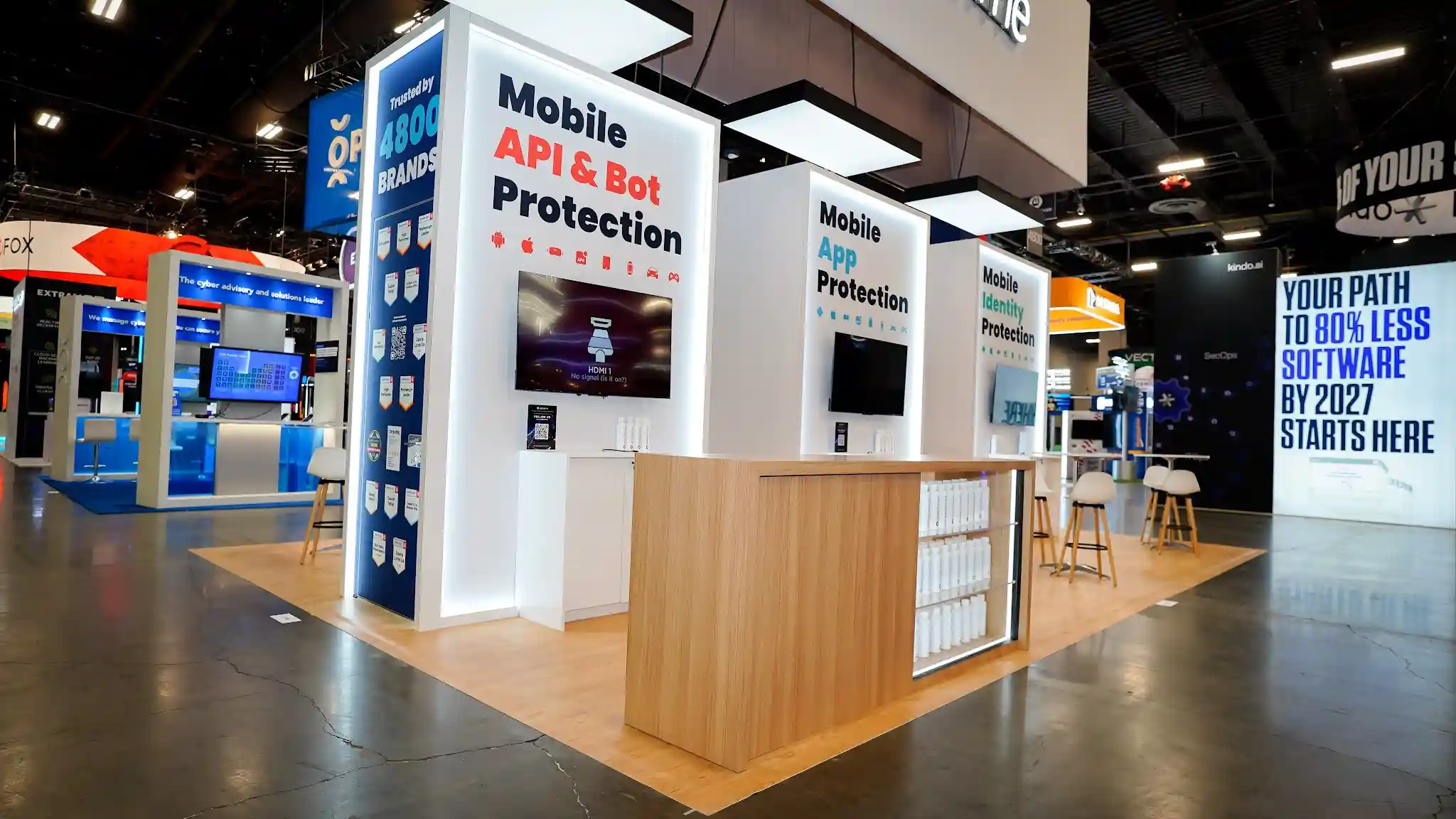 Three-quarter left-front oblique view of Appdome 20x30 island exhibit at Black Hat USA 2025 emphasizing the natural wood bar counter in foreground with open shelving and branded merchandise, three white product towers receding into background with LED edge lighting and flat-screen monitors, white overhead canopy with light panels illuminating the exhibit, raised wood platform flooring creating a defined footprint, neighboring Fox booth and 80 percent less software booth visible for context, and the overall hospitality-retail character of the Apple Store-inspired design at Booth 4746 Mandalay Bay Convention Center