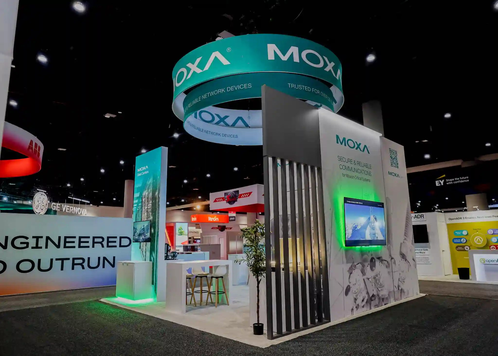 Moxa booth showing dark grey vertical architectural fins, green LED accent lighting behind monitors, recessed display with green halo effect, and meeting area with high-top stools