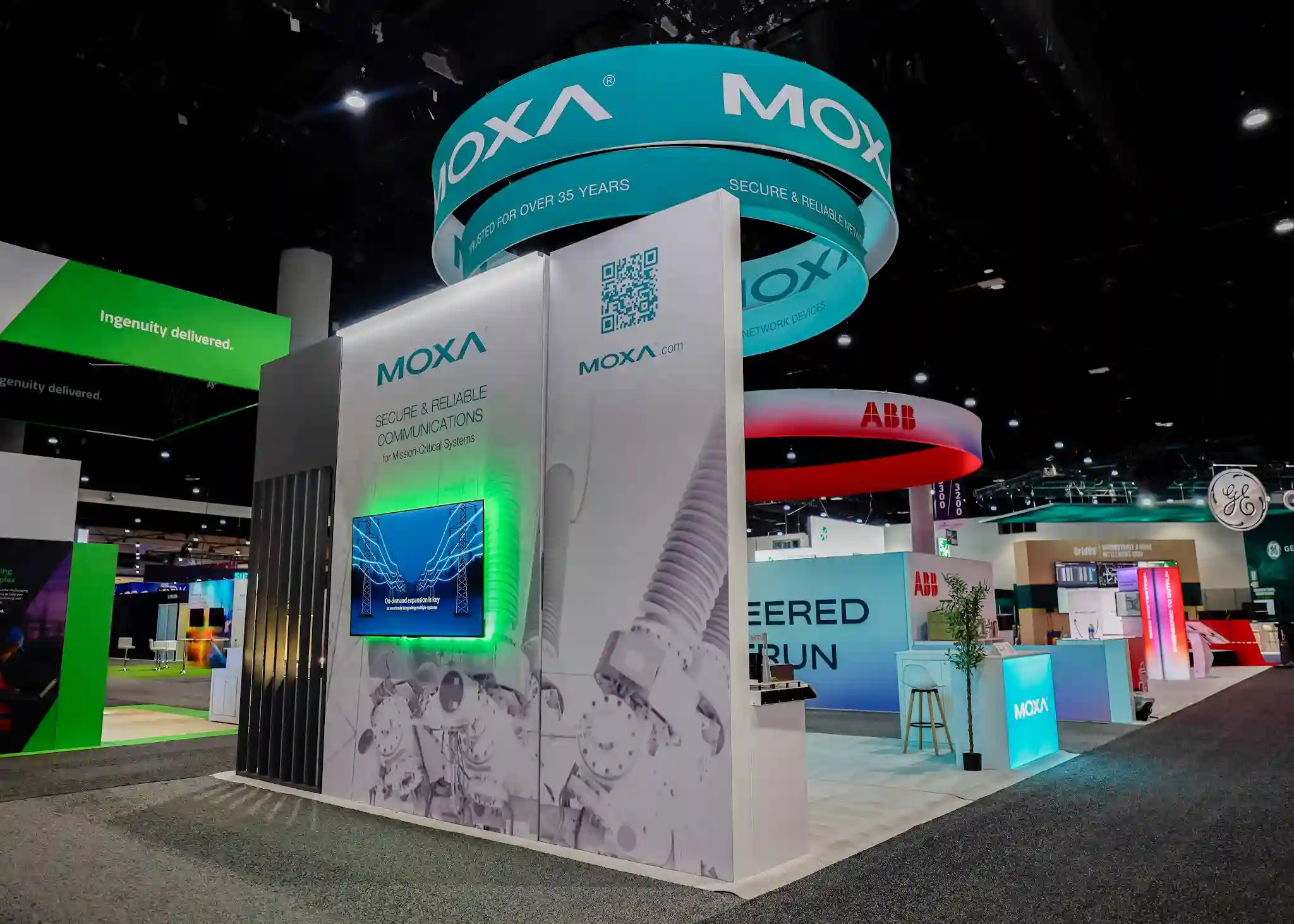 Multi-tiered circular hanging sign with Moxa logo in teal, Secure and Reliable Network Devices tagline, internally lit white panels, green LED backlighting on wall monitors below