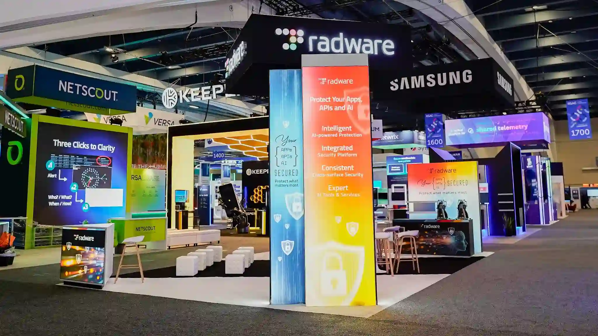 Wide panoramic establishing shot of the complete Radware 20x30 ft custom island exhibit at RSA Conference 2026 Booth S-1627 in the Moscone Center South Expo showing the full booth layout with the multicolor RGB LED gateway arch portal at center featuring a vivid red to orange to yellow gradient across the top bar and teal cyan vertical sides, a tall internally-illuminated multicolor gradient lightbox column displaying the Intelligent Integrated Consistent Expert value framework with shield and padlock icons on warm yellow-orange lower gradient, large rectangular matte black hanging overhead sign with luminous white radware lowercase wordmark and multicolor dot-grid icon suspended from ceiling rigging, white raised platform with cyan ice-blue LED strip underlighting creating a floating effect, white cube ottomans arranged in a semicircle in the aisle outside the booth boundary, central LED video wall visible through the arch showing animated network visualization, far-right demo counter with robotic prop, NETSCOUT teal booth visible at left, Keeper dark booth with honeycomb orange ceiling in center-left background, Samsung hanging signs at far right, RSAC conference floor decal at aisle boundary