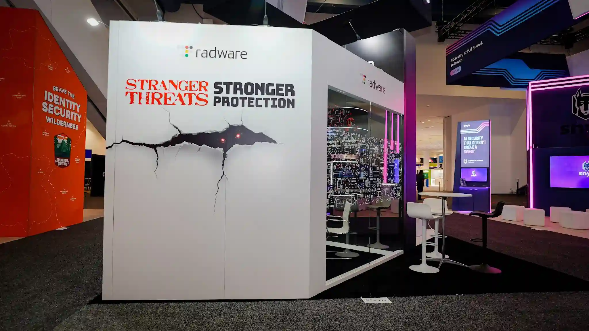 Close-up front view of the Radware booth primary exterior wall at RSA Conference 2025 showing the dramatic cracked-wall 3D graphic in full detail with white matte laminate surface appearing to fracture open from the center revealing glowing red monster creature eyes peering through the jagged tear, Stranger Threats in large red distressed Stranger Things-inspired horror typography above the crack, Stronger Protection in large bold black sans-serif below, radware colorful dot-cluster logo centered at top, cyan blue LED accent strip lighting along the base, dark charcoal carpet flooring, semi-visible interior meeting space on the right with dark walls featuring white cybersecurity icon line-art wallcovering, white tulip-base bar stools and round black pedestal bar tables inside, adjacent orange booth to the left reading Brave the Identity Security Wilderness, Snyk booth with purple neon lighting and AI Security at Full Speed No Sweat digital display visible far right, RSA Conference Moscone Center dark ceiling with overhead track lighting