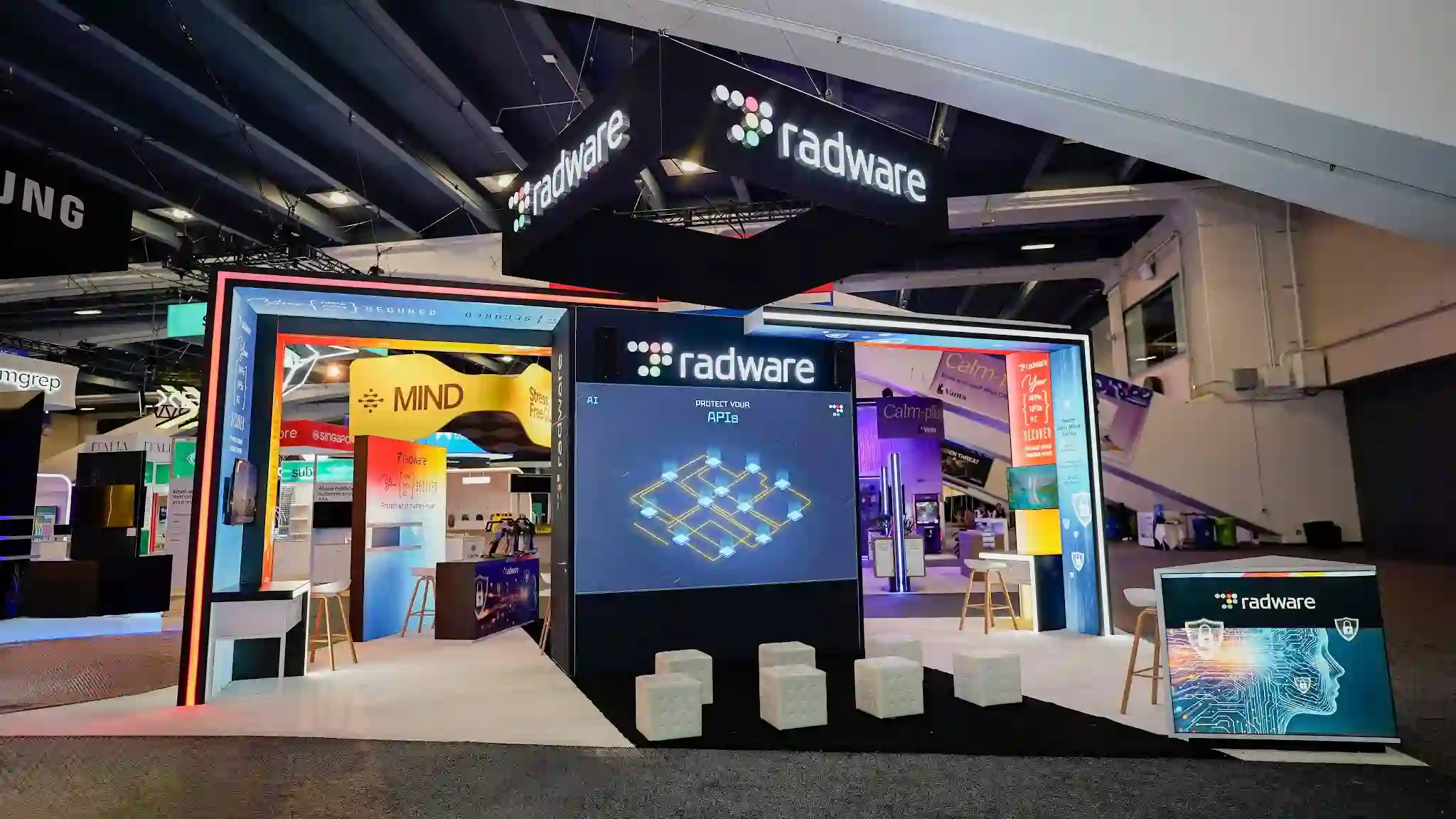 Interior view of the Radware booth at RSA Conference 2026 looking straight through the LED gateway arch from the aisle showing the complete U-shaped dual-wing interior layout with the large format LED video wall centered on a dark structural column displaying the radware logo AI subheading PROTECT YOUR APIs text and interconnected glowing gold lattice network visualization on deep navy blue, the left interior wing with vivid orange yellow and red graphic wrap panels creating a warm energetic atmosphere, the right interior wing with soft purple lavender and cool teal aqua panels creating a calm technical atmosphere, white cube ottomans arranged in a semicircle in front of the LED wall, bar-height bistro tables with round white tops and natural wood leg bar stools, multiple demo stations visible on both sides, dark navy cylindrical interior columns at corners, overhead hanging radware signs on both sides, white raised platform floor tiles throughout, neighboring Vanta teal booth visible through right exit, Keeper wolf icon background left, MIND bold yellow text on dark panel visible at far left