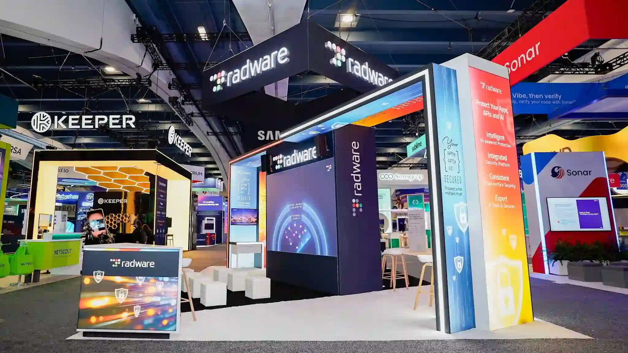 Front-right angle of the Radware RSA 2026 booth showing the LED gateway arch at center with the dark navy column displaying the multicolor Radware dot-grid logo, the tall multicolor gradient lightbox pillar prominently displaying Intelligent AI-powered Protection Integrated Security Platform Consistent Cross-surface Security Expert AI Tools and Services framework with shield padlock icons on warm yellow orange base, two overhead hanging radware signs visible simultaneously, Keeper dark blue booth with wolf icon in left background, Samsung hanging sign at far background, Sonar red sign at right background, white cube ottomans on platform