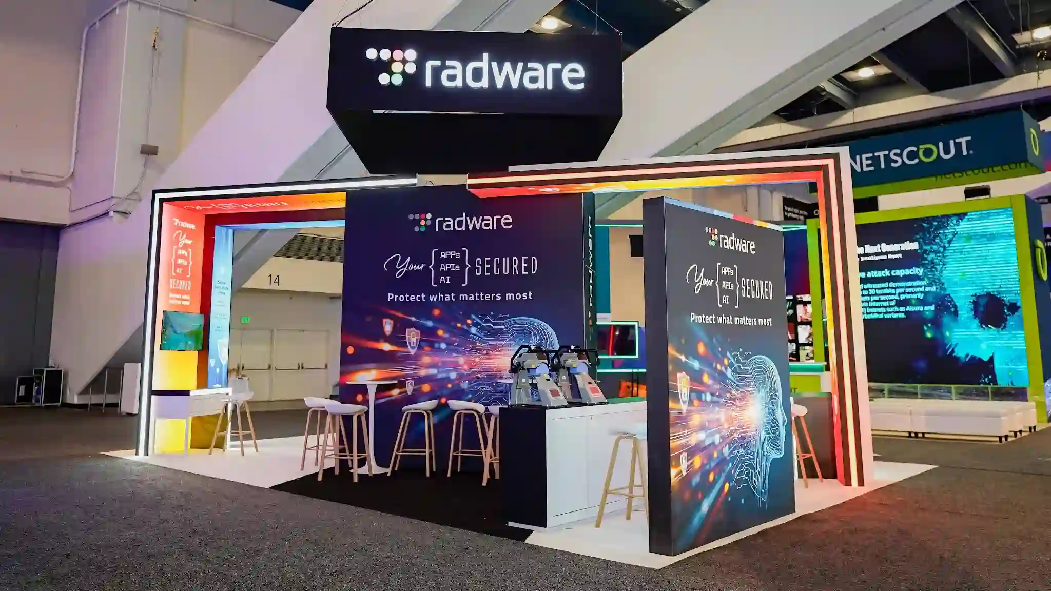 Front-facing hero shot of the Radware 20x30 ft custom island exhibit at RSA Conference 2026 Booth S-1627 taken from ground level centered on the main entrance showing the large rectangular matte black hanging overhead sign with luminous white radware lowercase wordmark and multicolor dot-grid icon dominating the upper frame, a central bar-height matte black demo counter with natural birch wood-tone bar stools in white, the multicolor RGB LED gateway arch portal visible in the booth interior with red to orange to yellow gradient on the top bar and teal sides framing the dark interior column, a tall white gradient lightbox column at right displaying the Your Apps APIs AI SECURED tagline and Agentic AI Security API Security Web Application Firewall Bot Management DDoS Protection Kubernetes WAAP product listing, white raised platform with cyan LED underlighting, freestanding demo kiosk at lower left with Radware branding and circuit board AI imagery, NETSCOUT teal hanging sign visible in far right background, Sonar red sign at far left background