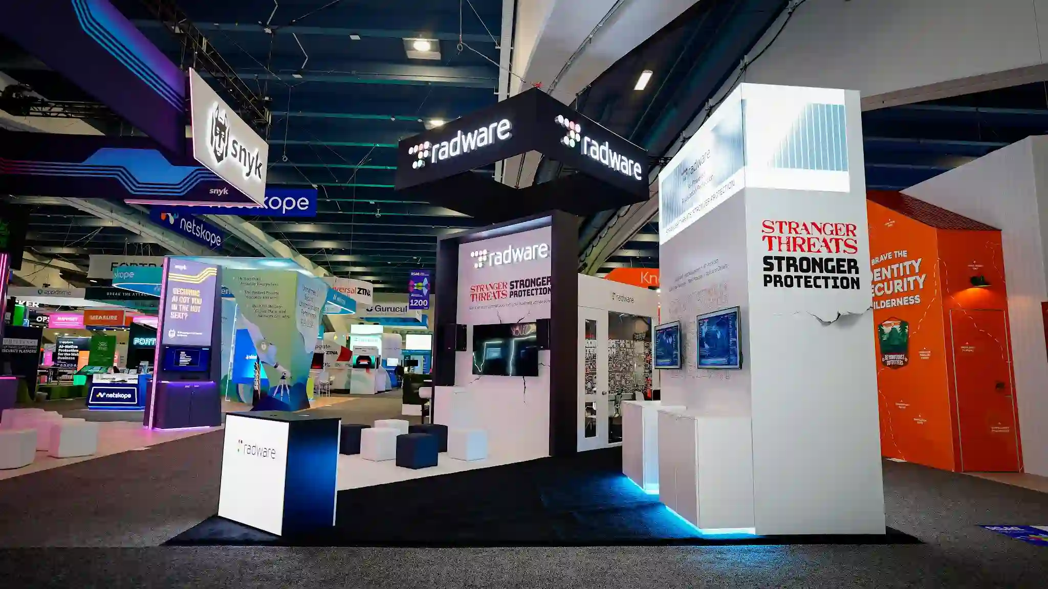 Wide-angle perspective view from the aisle of the Radware 20x30 island exhibit at RSA Conference 2025 showing the full booth configuration with tall white tower column on the right featuring Stranger Threats in red distressed Stranger Things-inspired typography and Stronger Protection in bold black text with dramatic cracked-wall graphic revealing glowing red monster eyes, large dark matte black overhead suspended canopy with radware logo in white on two visible sides, white reception demo podium with radware branding in the foreground left, dark navy blue cube ottomans clustered in open seating area inside the booth, interior flat-panel monitors displaying dark cybersecurity-themed graphics, electric cyan blue LED strip lighting along the base of the white tower structure, dark carpet flooring throughout, neighboring Snyk booth with purple navy branding and hanging banner visible upper left, Netskope blue-lit booth to the left, orange booth reading Brave the Identity Security Wilderness adjacent right, RSA Conference Moscone Center show floor with dark ceiling and industrial track lighting