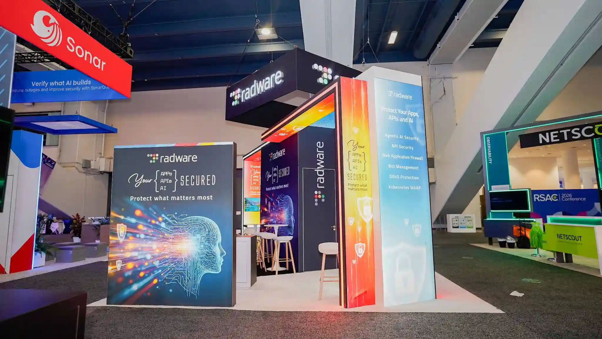 45-degree front-left angle shot of the Radware booth at RSA Conference 2026 showing the multicolor RGB LED gateway arch portal in full chromatic detail with the top horizontal bar transitioning from vivid red through warm orange to golden yellow and both vertical sides in teal cyan, the large rectangular matte black hanging overhead sign with luminous white radware wordmark and multicolor dot-grid icon centered above, a tall dark navy column at left of arch displaying the Radware logo with multicolored dot icon, the tall multicolor gradient lightbox pillar at right transitioning from sky blue at top through warm yellow to safety orange at bottom with the Your Apps APIs AI SECURED tagline and shield padlock icons, white raised platform with cyan LED underlighting visible at base, interior through arch showing warm golden lighting and animated LED video wall content, Sonar red booth visible at top right background