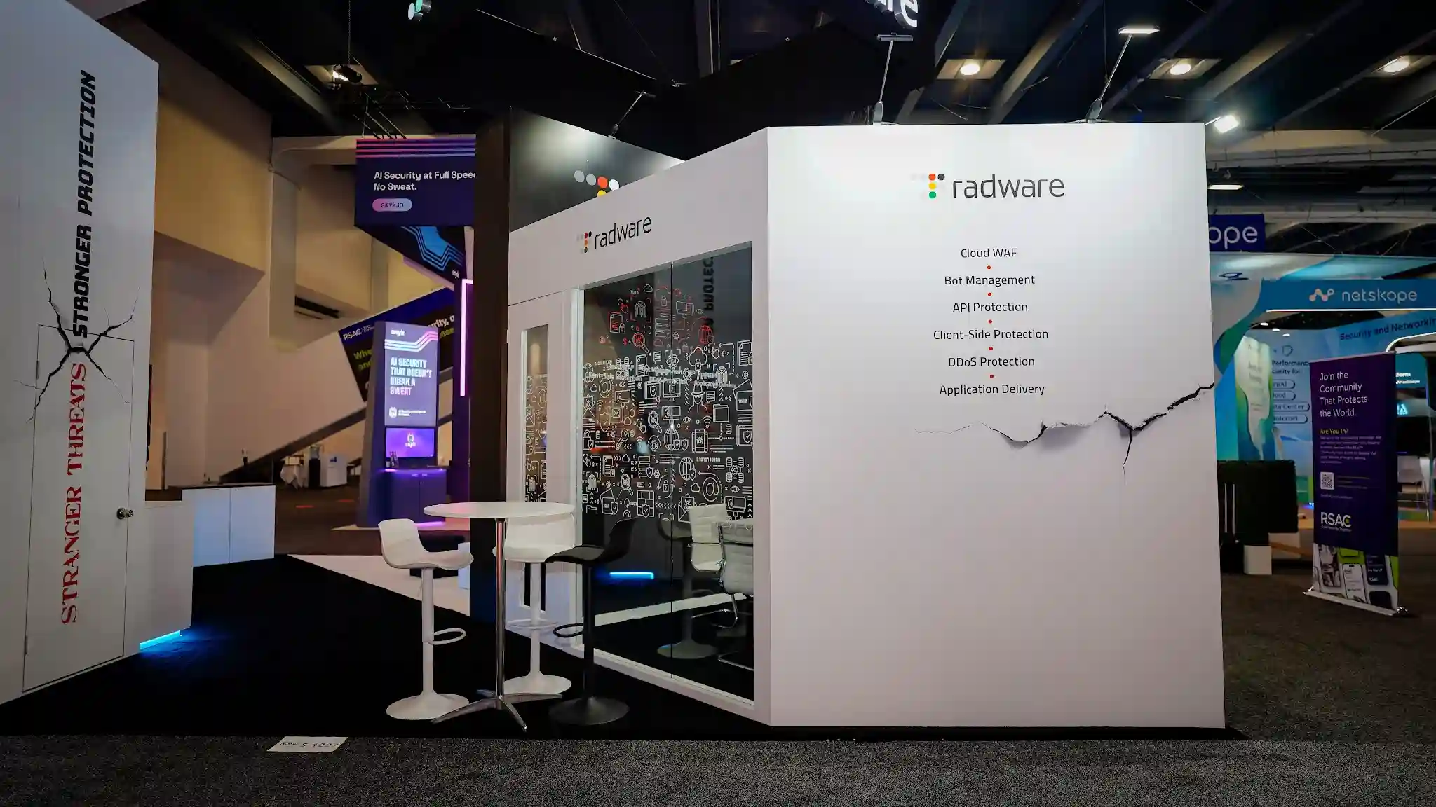 Straight-on aisle view of the Radware booth right side at RSA Conference 2025 showing the enclosed glass-walled meeting room structure with dark interior walls featuring dense white cybersecurity icon line-art wallcovering depicting servers shields locks cloud elements and network diagrams on black background, transparent glass front panels revealing interior monitors displaying Radware-branded blue teal technology map, radware colorful dot-cluster logo on the upper white exterior, right outer white wall panel displaying clean product solution list in black text including Cloud WAF Bot Management API Protection Client-Side Protection DDoS Protection Application Delivery with cracked-wall graphic motif extending into the panel, left narrow tower column partially visible with Stranger Threats Stronger Protection vertical signage in red and black, two modern white tulip-style pedestal bar stools with chrome-effect bases paired with round white bar-height table positioned outside the booth for informal demos, cyan blue LED underglow creating a cool floating effect at the base, dark charcoal carpet flooring, neighboring Netskope booth with teal blue branding and RSAC conference signage to the right, Snyk purple backlit booth with AI Security at Full Speed No Sweat messaging visible in background center, Moscone Center dark ceiling with overhead track lighting