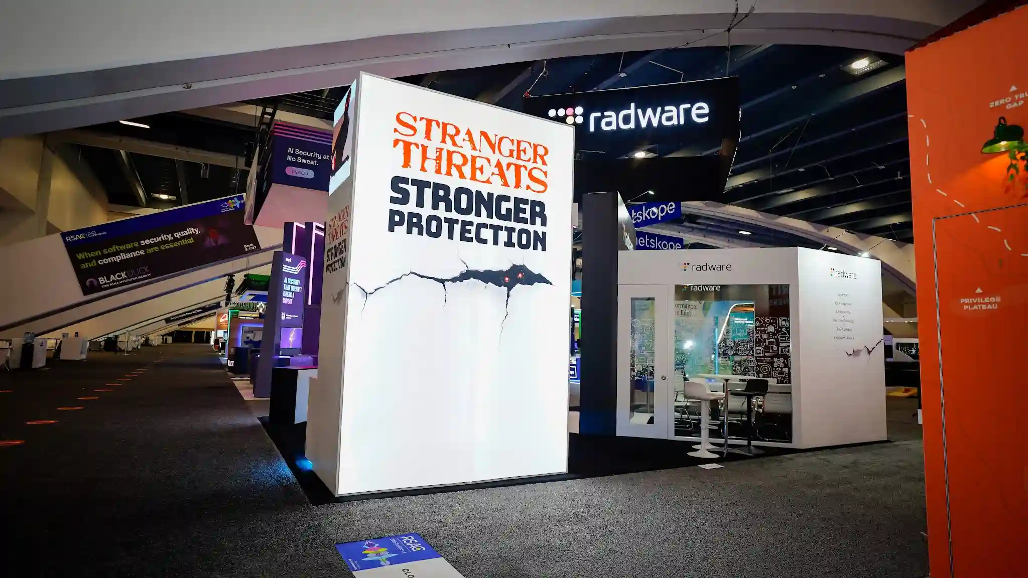 Wide aisle perspective view of the Radware backlit SEG lightbox tower at RSA Conference 2025 showing the brilliantly illuminated white panel glowing with internal LED backlighting, Stranger Threats in large orange-red distressed Stranger Things-inspired typography at top, Stronger Protection in bold dark sans-serif below, dramatic cracked-wall graphic with glowing red creature eyes rendered on translucent stretch fabric creating an almost supernatural luminous effect, cyan blue LED accent strip at the base, the main Radware enclosed meeting room structure visible in the background with dark overhead canopy showing radware logo in white, product list panel partially visible on the right showing Cloud WAF Bot Management API Protection, white tulip-base bar stools and round bar table visible inside the booth through glass panels, neighboring Netskope booth with teal hanging banners center-background, orange-branded booth with vintage industrial pendant lamp far right, Snyk and Black Duck booths partially visible far left, RSA Conference badge pamphlet on the dark show floor in foreground, Moscone Center dark ceiling with industrial rigging overhead
