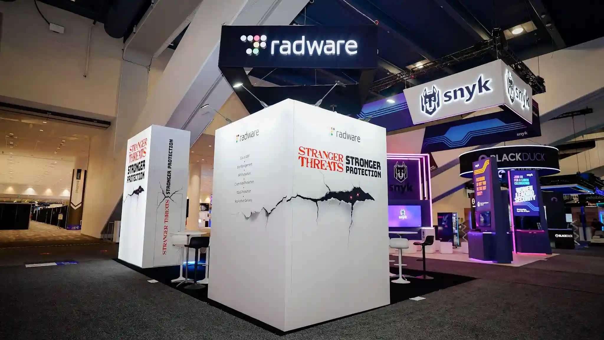 Wide-angle overhead view of the complete Radware 20x30 custom island exhibit at RSA Conference 2025 Booth S-1227 showing three distinct white structural elements against dark show floor carpet: a tall narrow backlit SEG lightbox tower on the left displaying Stranger Threats Stronger Protection campaign messaging in red distressed and bold black typography with cracked-wall graphic and glowing red bat-like creature eyes, a large central enclosed white cube structure housing the glass-walled meeting room with dark interior walls visible through transparent panels, a dark matte black suspended overhead canopy with radware logo in white and colorful dot-cluster branding, cyan blue LED accent strip lighting at the base of all white structures, product feature list panel showing Cloud WAF Bot Management API Protection Client-Side Protection DDoS Protection Application Delivery, white round bar-height tables and pedestal-base bar stools inside the booth, neighboring Snyk booth with purple neon lighting and suspended white banners to the right, Black Duck dark navy circular kiosk visible in background, RSA Conference Moscone Center dark ceiling with industrial rigging and track lighting overhead
