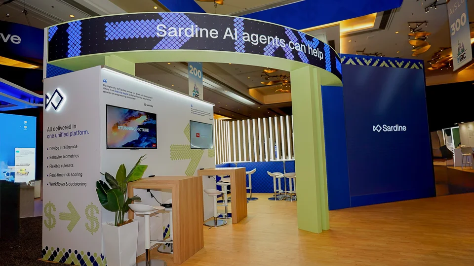 Interior view shot from inside the Sardine booth looking outward through the large curved sage-green entry archway approximately 12-14 feet tall with smooth rounded columns and top beam, curved horizontal canopy above reading Sardine AI agents can help in bold white on dark navy with sparkling dot diamond mosaic patterns in yellow and teal, navy-blue back wall with Sardine wordmark and logo in white and subtle pixel-dot texture, left interior wall with printed content panels including testimonial quotes diagrams and five feature bullets for unified platform, two flat-panel monitors showing product demos, vertical wooden slat walls with built-in warm LED strip lighting, potted tropical plant near archway column, tall light wood poseur table in center, white chrome-base bar stools, light wood-grain LVT plank flooring throughout interior