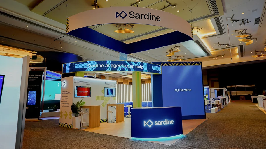 Definitive front-on hero shot of the complete Sardine AI Agents 20x20 ft custom island booth at MRC Vegas 2026 Booth 217 at ARIA Resort and Casino Las Vegas showing the large sweeping curved white-and-blue ribbon hanging sign approximately 20 feet wide displaying Sardine wordmark with angular diamond logo in bold dark navy on white face with blue underside and yellow-black chevron accents at ends, left center zone with tall tower section featuring horizontal LED ticker banner reading Sardine AI agents can help in white on royal blue with sparkling pixel diamond graphic accents and two product demo monitors mounted inside showing platform interfaces, right zone with large standalone navy-blue billboard wall panel bearing centered Sardine wordmark and logo in white, prominent curved reception desk at center front finished in royal blue with sardine wordmark and logo in white and vivid teal cyan LED strip at base creating glowing halo on light honey-toned wood-grain LVT plank flooring, white chrome-base bar stools throughout, light wood poseur tables, open airy interior layout, neighboring dark fintech identity verification booth visible to left, dark gray convention hall carpet surrounding booth