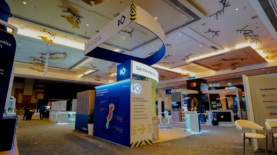 Wide-angle low-vantage perspective of the Sardine AI custom island trade show booth at MRC Vegas 2026 emphasizing the dramatic curved ribbon-shaped overhead hanging sign with pure white outer face and vivid royal-blue underside reading Agentic Platform to Fight Fraud and Abuse with Sardine angular diamond logo icon in blue and yellow-and-black diagonal chevron warning-stripe graphics at terminal ends, large solid wall panels in deep royal blue with subtle pixel dot-grid texture, column section facing main aisle reading SARDINE.AI, white curved reception counter with teal LED strip base, smartphone graphic with heat-map visualization for bot-driven carding attack detection, five-bullet unified platform list including device intelligence behavior biometrics flexible rulesets real-time risk scoring workflows and decisioning, ARIA Resort ballroom venue with coffered ceilings recessed can lighting amber chandeliers and decorative gold sculptural fish figures, neighboring Southwest Airlines Nu Skin Autotrader Wyndham Hotels Mastercard booths visible in background
