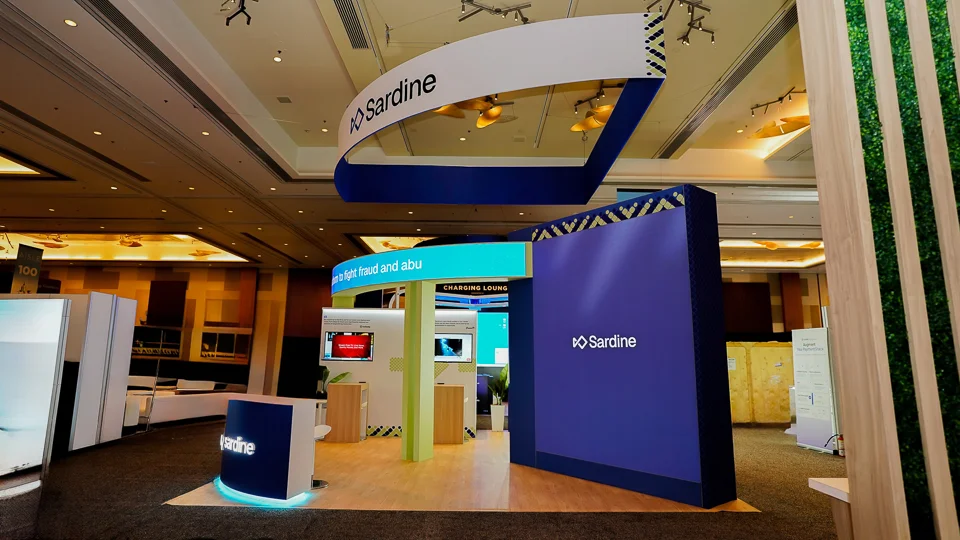Corner-angle view of the Sardine booth showing right-side large navy-blue billboard panel with Sardine wordmark and yellow-black chevron accent, overhead ribbon hanging sign, LED ticker band, Charging Lounge in teal text, interior demo monitors, curved reception counter, wood flooring, neighboring exhibitors with plant walls visible