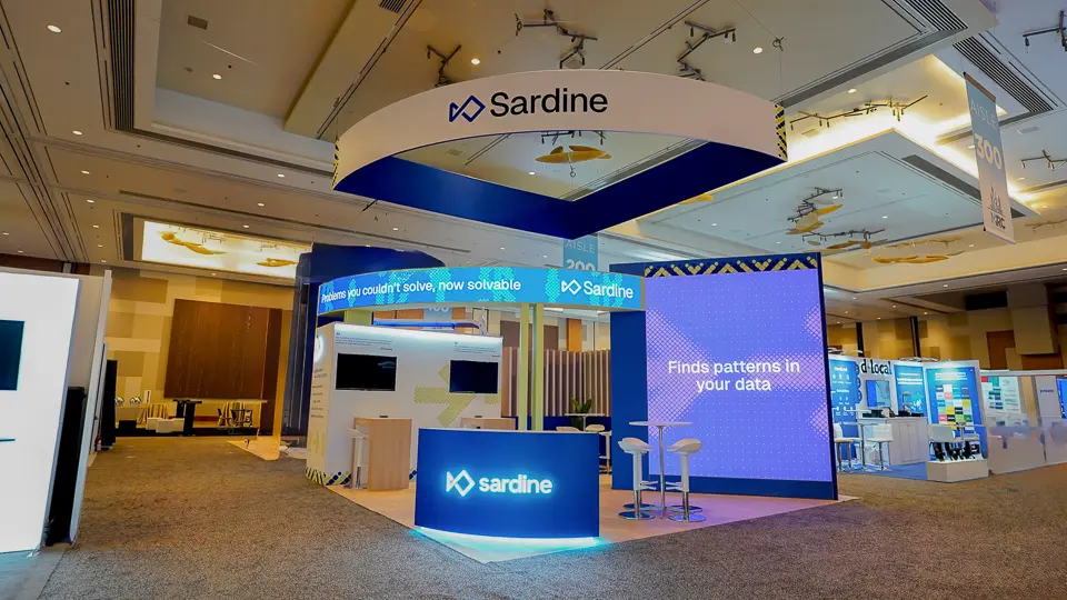 Clean centered head-on view of the full Sardine 20x20 ft island booth at MRC Vegas 2026 showing the complete booth structure with overhead curved white-and-blue ribbon hanging sign displaying Sardine wordmark and logo with yellow-black chevron accents, right side large freestanding LED video wall displaying purple-blue gradient with Finds patterns in your data text in white, left side multi-story tower with horizontal LED ticker banner scrolling Problems you couldnt solve now solvable in white on electric blue, flat-panel monitor on light wood slat-panel wall backdrop showing software interface, vertical wooden blade slat elements, white chrome-base bar stools, tall light wood poseur counter table inside, curved white reception desk with blue LED base at front, light wood-grain LVT flooring within booth, dark gray carpet surrounding, d/local fintech booth visible on right, Aisle 300 ceiling sign