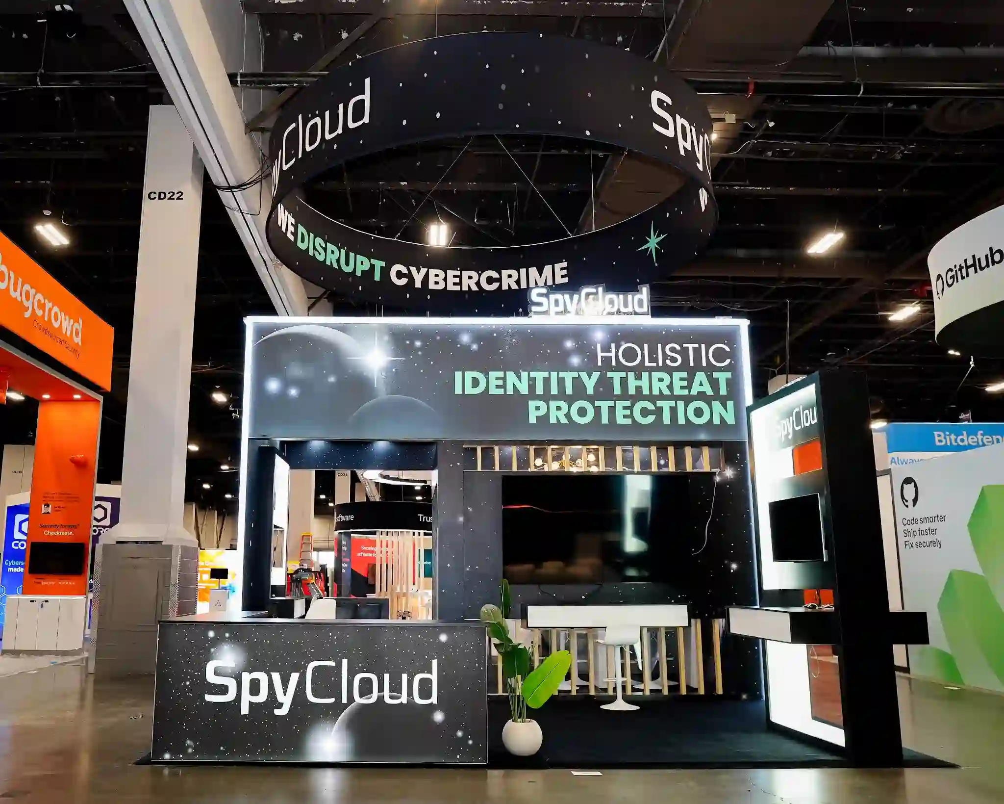 Front view of SpyCloud 20x20 custom island exhibit at Black Hat USA 2025 showing dark cosmic starfield graphic panels with teal accent text reading Holistic Identity Threat Protection, large circular overhead hanging sign with SpyCloud logo and We Disrupt Cybercrime messaging, bright white LED perimeter lighting framing the entire booth structure, branded reception counter at center front, white bar stools, and green tropical plant decoration at Booth 4821 in the Mandalay Bay Convention Center Business Hall
