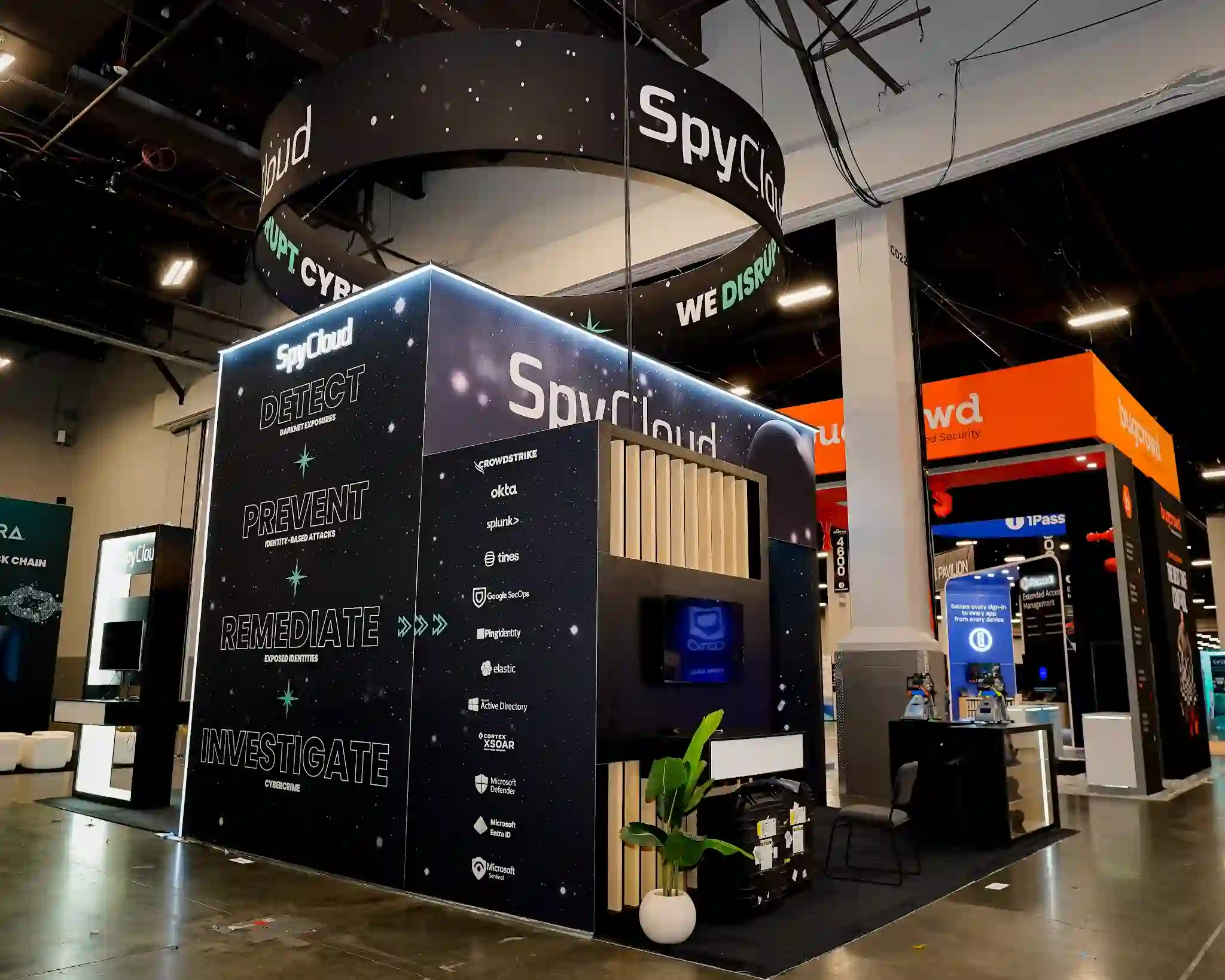 Corner side angle view of SpyCloud island exhibit at Black Hat USA 2025 emphasizing the large circular backlit hanging sign with SpyCloud logo and We Disrupt Cybercrime teal ring text, left exterior panel showing Detect Darknet Exposures Prevent Identity-Based Attacks Remediate Exposed Identities Investigate Cybercrime workflow sequence, right panel displaying technology partner integration logos including CrowdStrike Okta Splunk Tines Google SecOps Ping Identity Elastic Active Directory Cortex XSOAR Microsoft Defender Microsoft Entra ID Microsoft Sentinel, white LED edge lighting framing entire structure, dark cosmic starfield graphic panels throughout