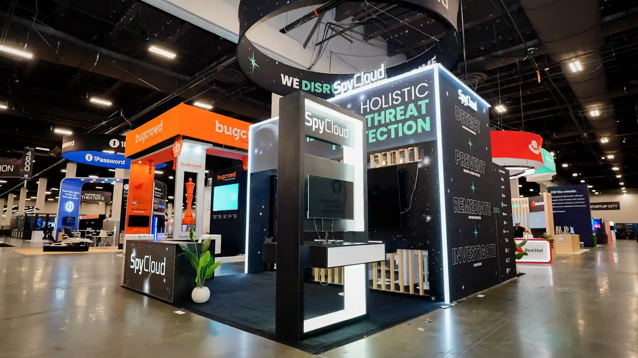 Side corner angle view of SpyCloud 20x20 island exhibit at Black Hat USA 2025 showing two exterior walls simultaneously with dark cosmic starfield panels, bright white LED edge trim on all structural edges, right-side wall displaying sequential workflow text reading Detect Darknet Exposures Prevent Identity-Based Attacks Remediate Exposed Identities Investigate Cybercrime in teal and white, front reception counter with SpyCloud branding, green tropical plant in white pot, and neighboring Bugcrowd and 1Password booths visible in background