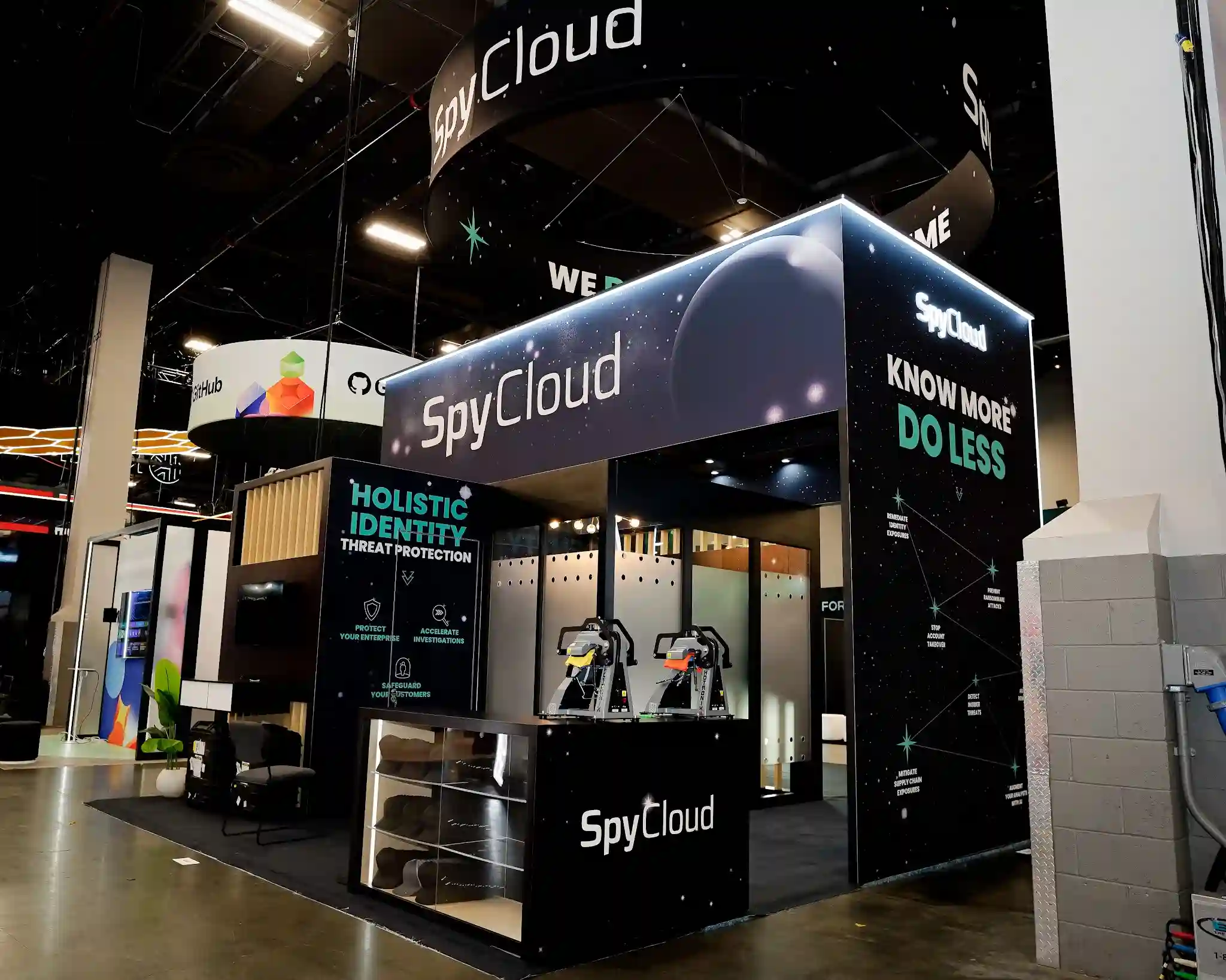 Front-left corner perspective of SpyCloud 20x20 island exhibit at Black Hat USA 2025 showing two exterior faces with dark cosmic starfield graphics, left face reading Holistic Identity Threat Protection with sub-bullets Protect Your Enterprise Accelerate Investigations Safeguard Your Customers, right face reading Know More Do Less in teal and white text, SpyCloud circular hanging sign overhead, natural wood slat panel visible through entry, multiple flatscreen monitors inside on stands, dark reception counter with SpyCloud logo at center front, LED perimeter lighting, and neighboring GitHub booth visible in background