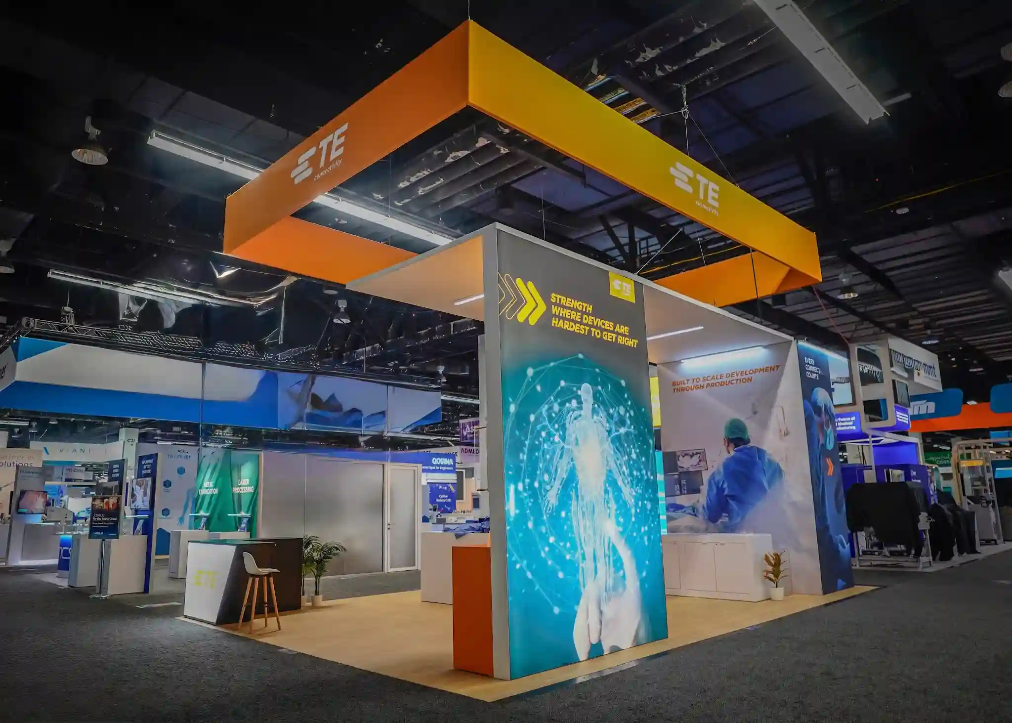 Large square orange-framed hanging sign with white TE Connectivity logo on all sides, cyan-blue backlit digital human skeleton wall below, Strength Where Devices Are Hardest to Get Right messaging, and wood-grain booth flooring visible