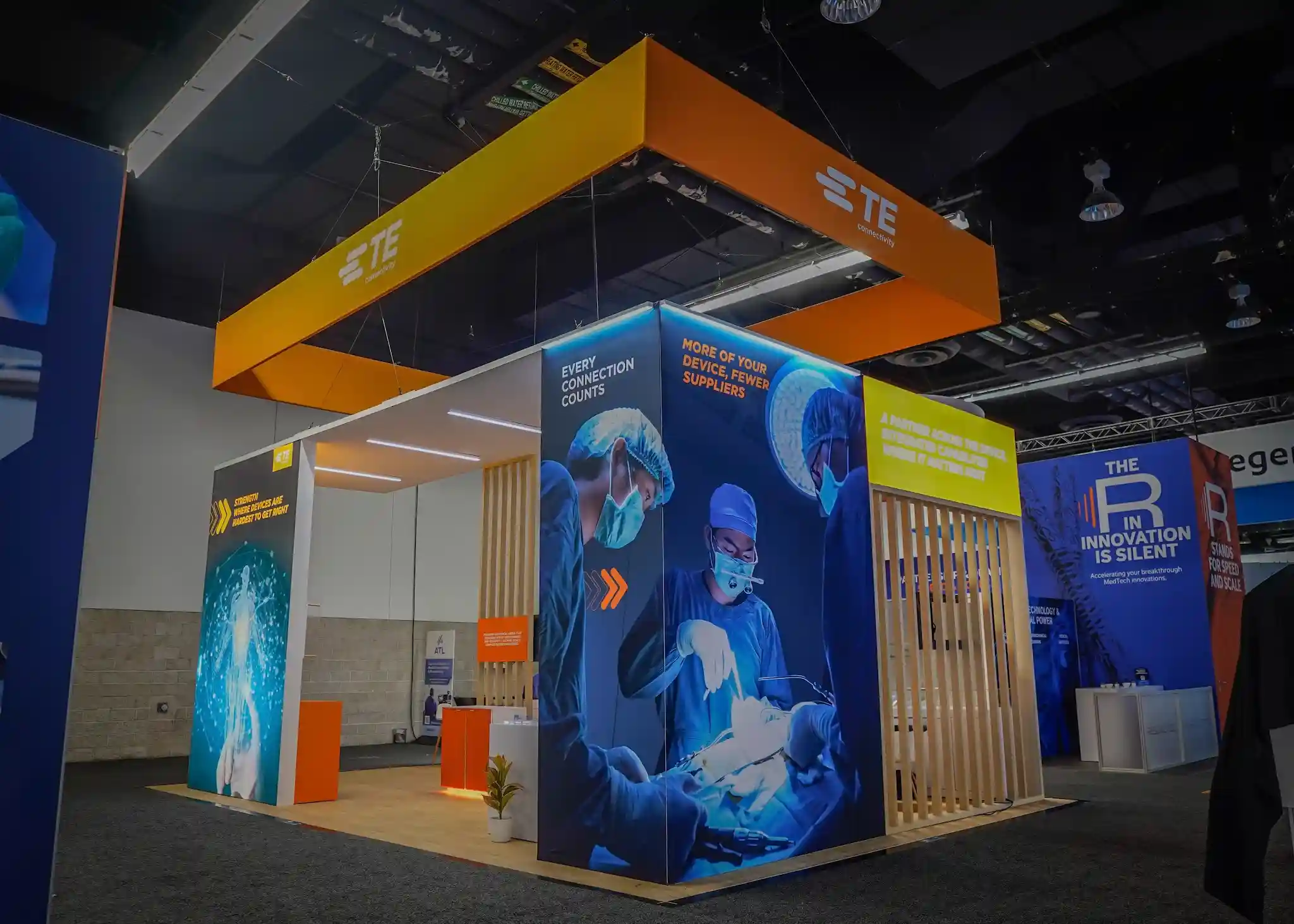 TE Connectivity booth corner featuring large-scale fabric graphics depicting a surgical environment, Every Connection Counts and More Of Your Device Fewer Suppliers messaging, overhead LED light bars, and vertical wooden slat wall with texture detail