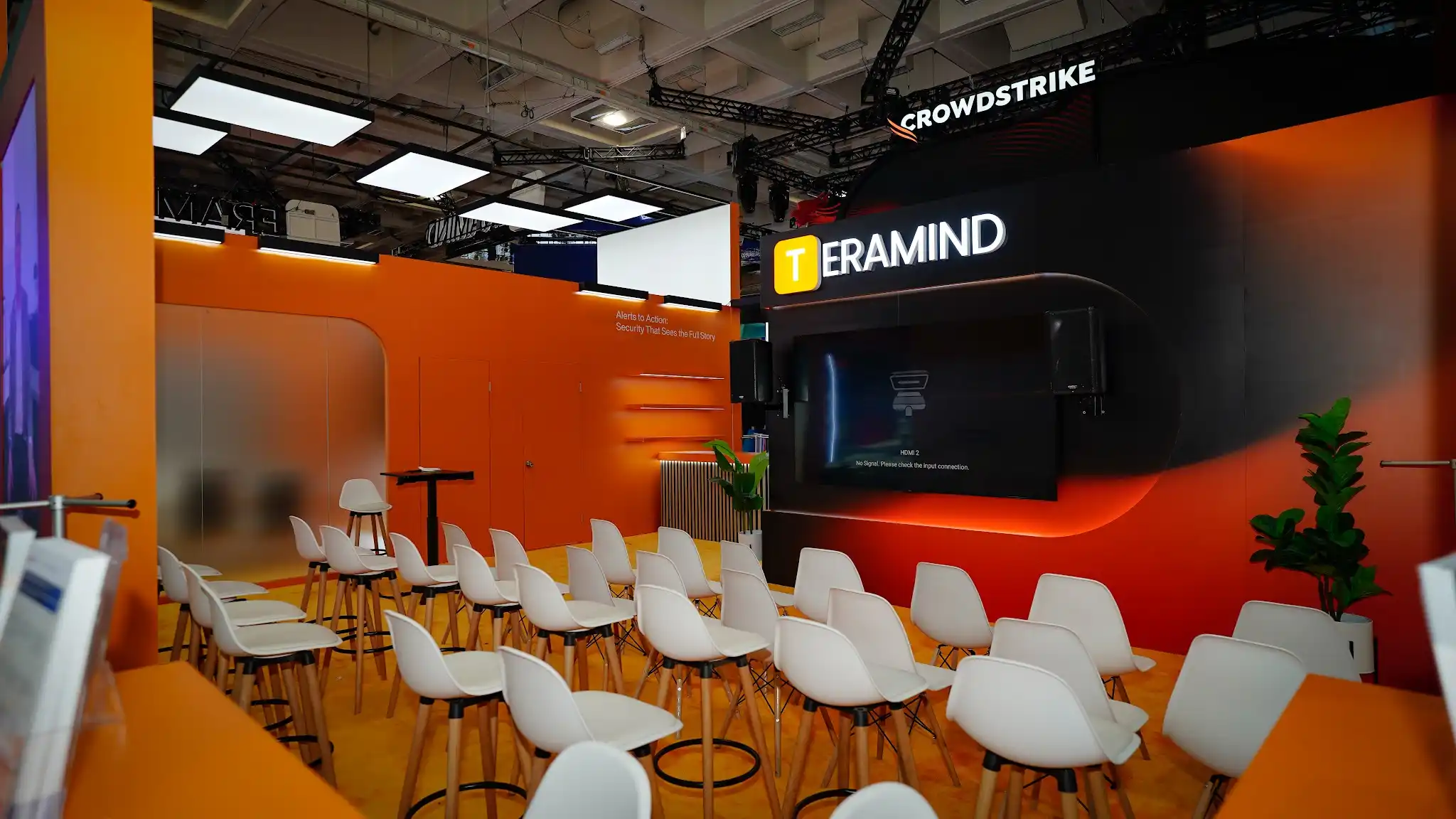 Interior view of the Teramind in-booth presentation theater at RSA Conference 2025 showing approximately 25 to 30 white chairs arranged in theater-style rows facing a large 75 to 85 inch presentation screen mounted on the dark charcoal back wall, blonde wood slat columns flanking the theater entrance creating a warm organic frame, burnt-orange accent lighting and panels surrounding the screen area, TERAMIND branding and product messaging visible on side panels including Teramind Insights See What Others Miss With AI-Powered Security Intelligence, LED ceiling panel tiles providing bright overhead illumination, amber carpet flooring throughout the theater zone, CrowdStrike booth with red branding and large overhead signage visible in the background through the open side of the theater, RSA Conference attendees seated in the presentation area