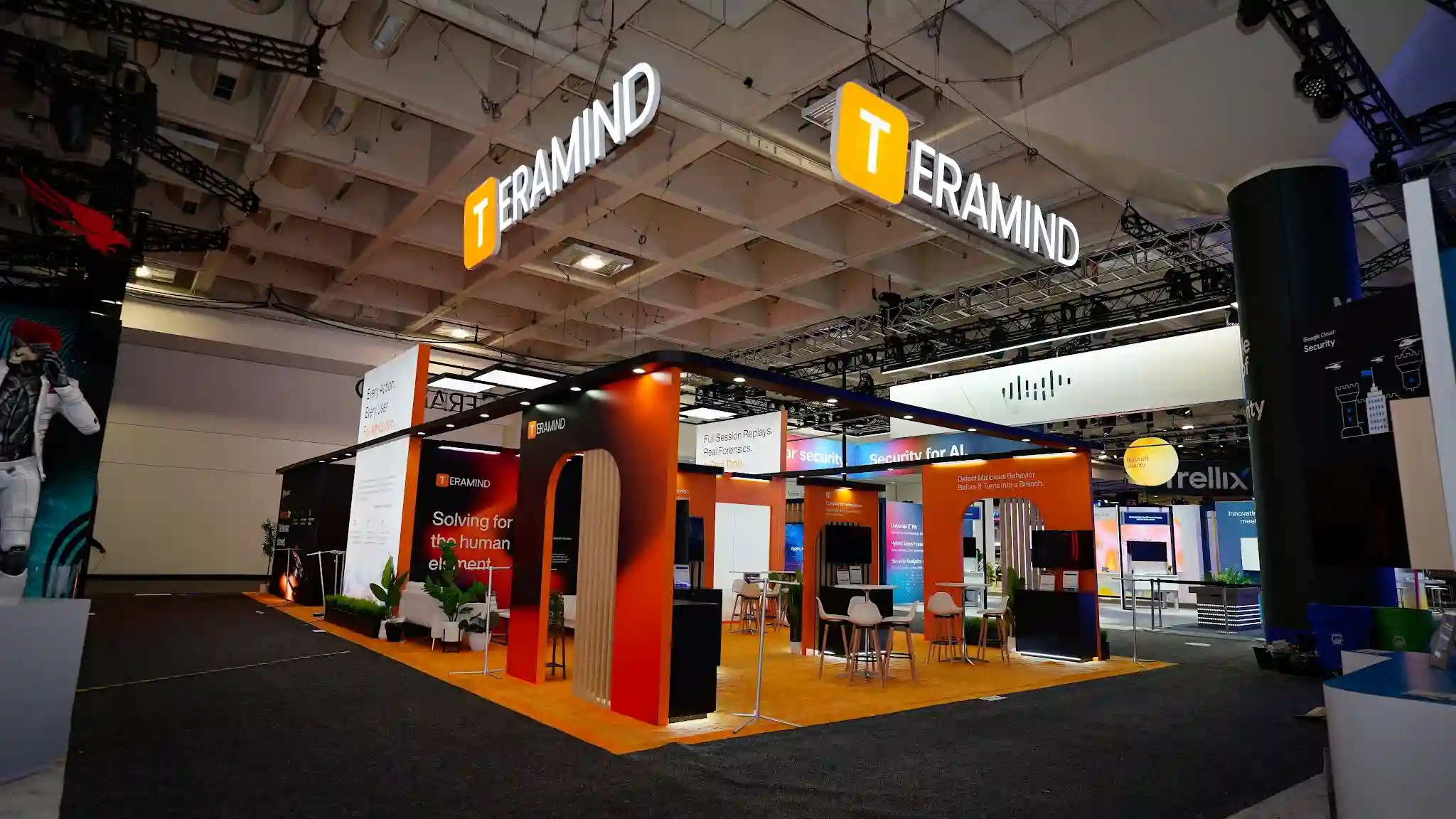 Close exterior view of the Teramind booth showing high-top networking area with white round tables and Eames-style chairs, burnt-orange and charcoal exterior panels, TERAMIND overhead signage, CrowdStrike red-branded booth clearly visible adjacent to the right, RSA Conference badge-wearing attendees in foreground