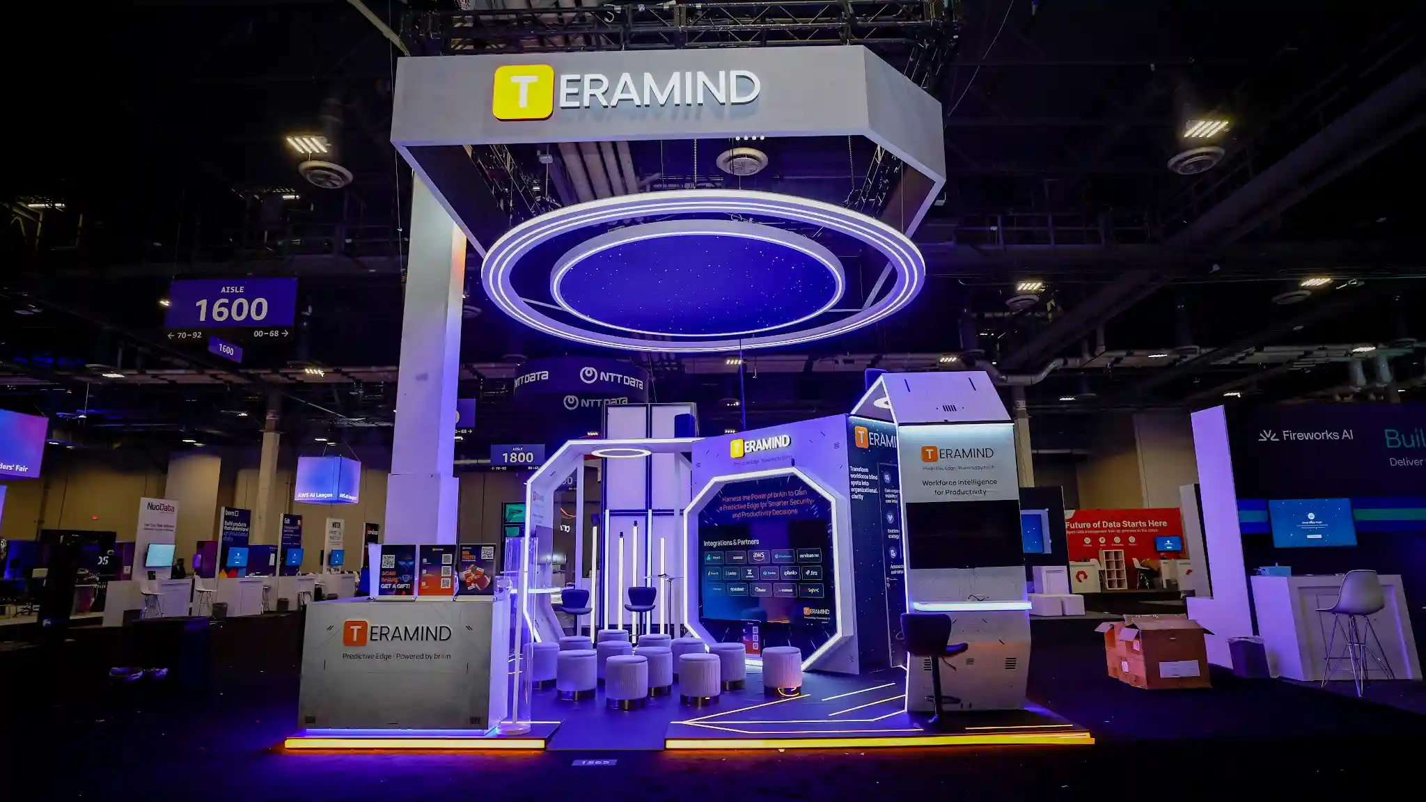 Wide-angle view of Teramind booth showing massive overhead sign, concentric blue LED halo rings, reception counter, demo kiosks, and yellow LED floor perimeter lighting