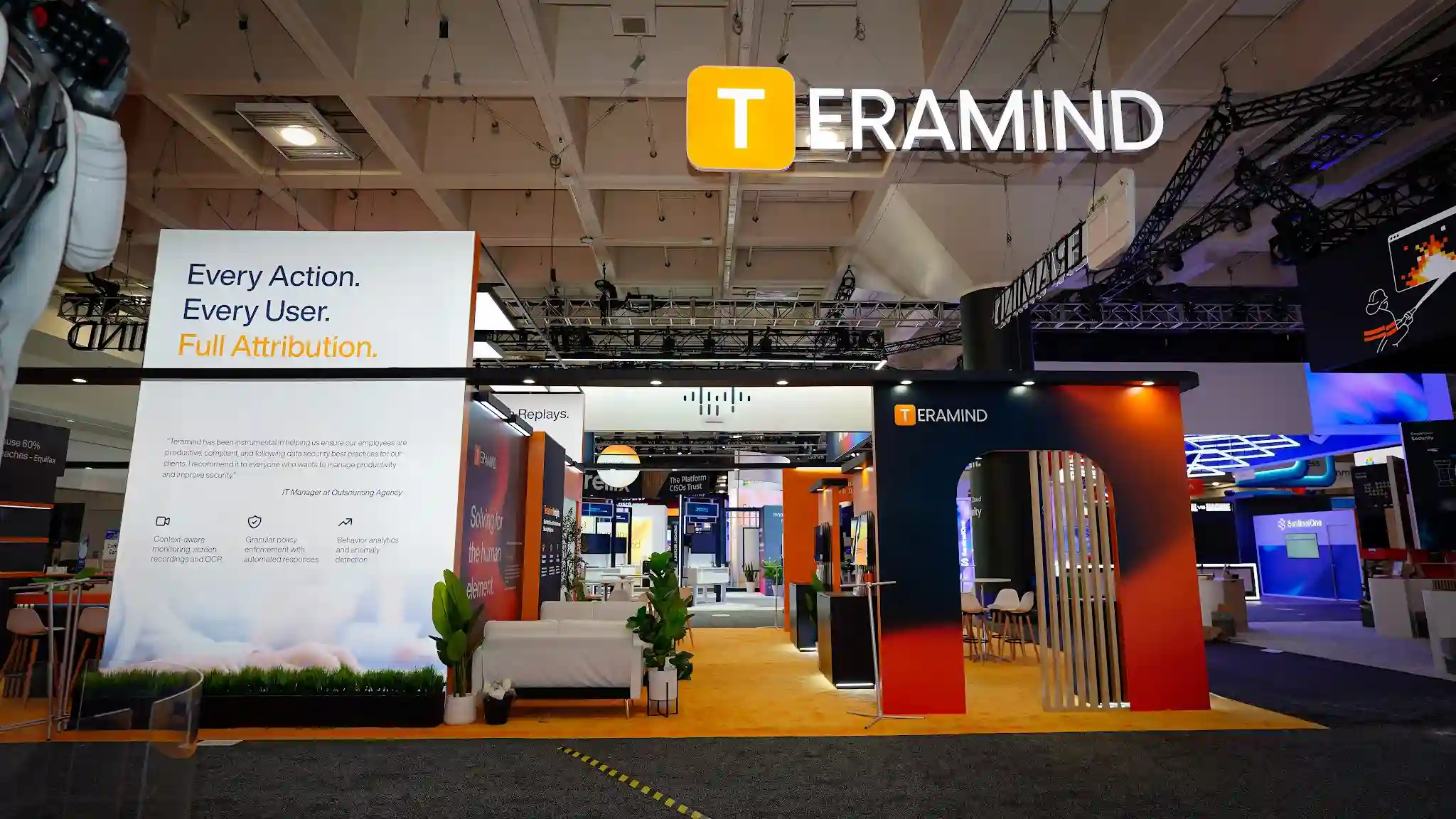 Theater entrance and demo kiosk row of the Teramind booth at RSA Conference 2025 showing the dedicated presentation theater entrance flanked by wood slat columns, row of product demo kiosk monitors on the right displaying Teramind platform interfaces, burnt-orange accent panels with product messaging, amber carpet flooring, LED ceiling lighting