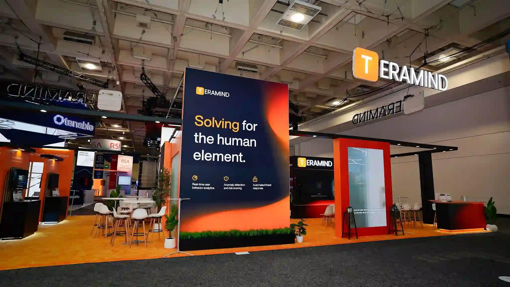 Exterior corner perspective of the Teramind 30x60 ft Diamond Sponsor island booth at RSA Conference 2025 from a different aisle angle showing the theater entrance and lounge area visible through the booth structure, blonde wood slat architectural elements framing the open sides, deep charcoal overhead canopy with TERAMIND three-dimensional logo sign and golden-T icon suspended above, burnt-orange accent panels and signage with Solving for the human element tagline, white low-profile sofa seating in the lounge zone with round coffee table, tropical green plants including ferns and olive tree adding organic warmth to the space, amber carpet flooring throughout, LED ceiling panel tiles creating bright even illumination, white Eames-shell chairs visible at networking tables, Moscone Center North Hall convention floor with neighboring cybersecurity vendor booths in background