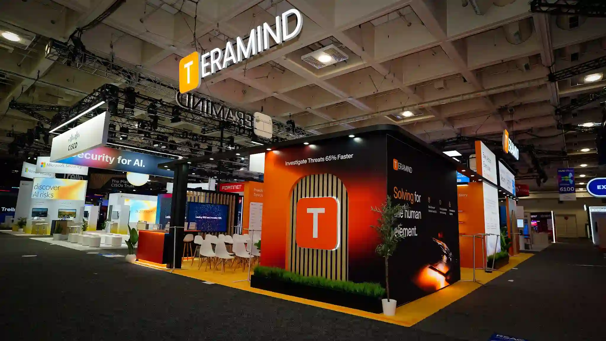 Aisle-side perspective view of the Teramind 30x60 ft island booth at RSA Conference 2025 showing the burnt-orange and deep charcoal exterior fascia with TERAMIND branding and golden-T logo, blonde wood slat architectural detail visible at the entrance, large overhead three-dimensional TERAMIND sign suspended from the convention ceiling against dark rigging, product messaging panels displaying key value propositions including Solving for the human element in orange and white typography, amber carpet flooring visible at the booth perimeter, white high-top networking tables and Eames-style chairs in the foreground, neighboring Google Cloud Security and Cisco booth signage visible in the background, Tenable dark booth visible across the aisle, RSA Conference Moscone Center North Hall environment with dark ceiling and overhead track lighting, attendees walking the aisles between Diamond Sponsor exhibits