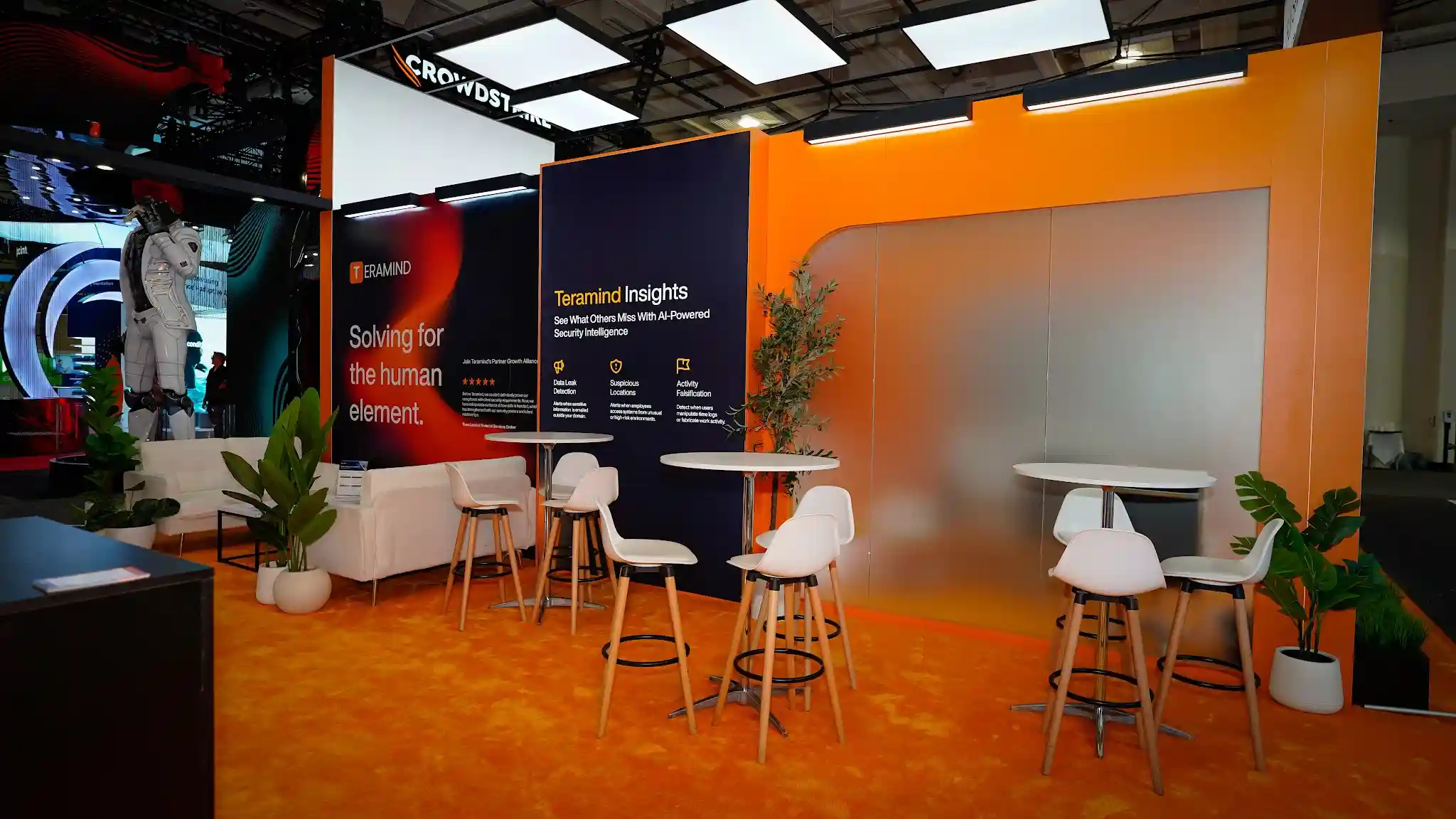 Interior lounge and meeting area of the Teramind booth showing white low-profile sofa seating with round coffee table, private meeting alcove with frosted glass walls visible in the background, tropical plants and greenery adding organic warmth, burnt-orange accent panels, amber carpet, neighboring CrowdStrike booth with distinctive red branding visible through the open side of the booth