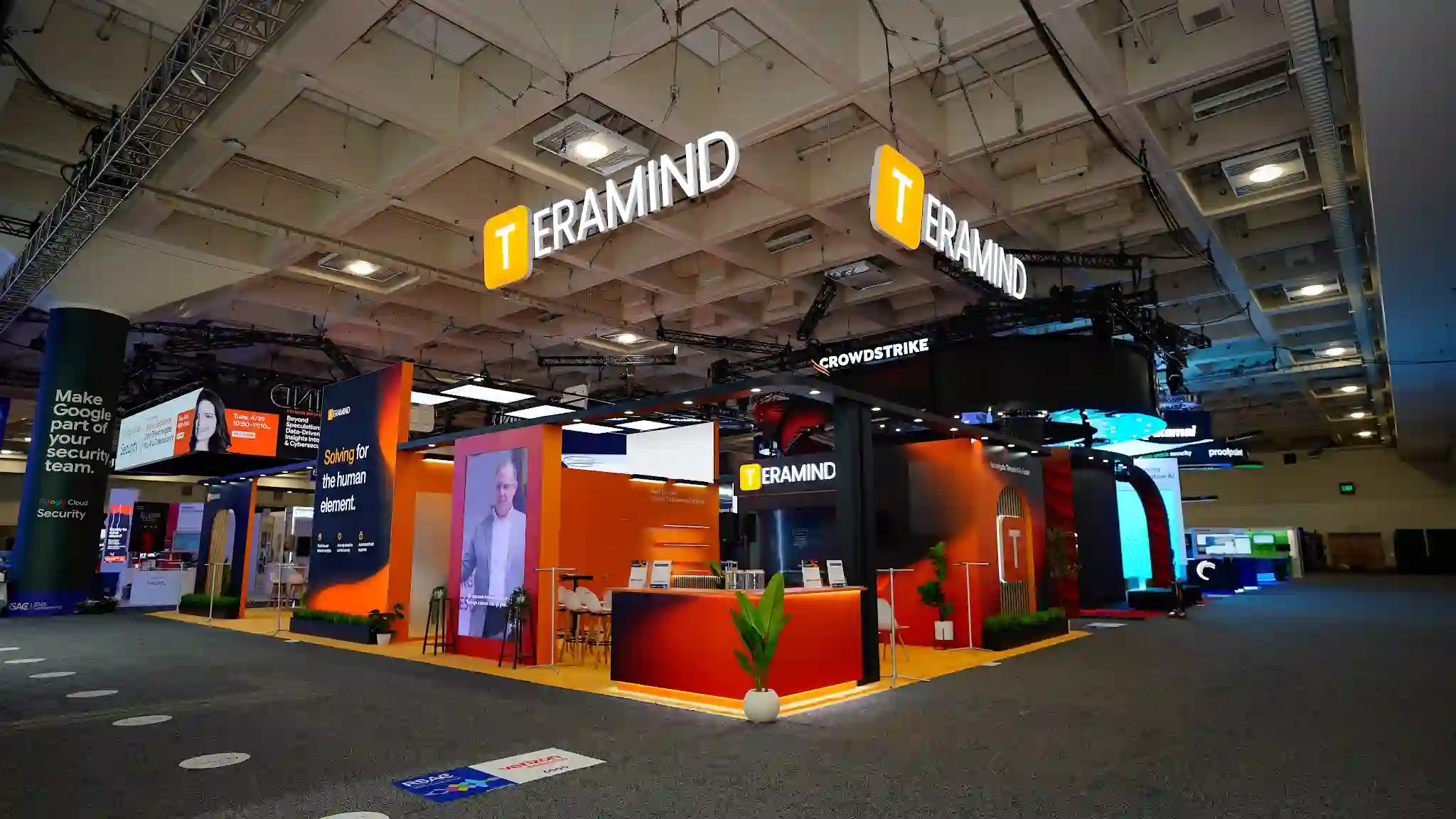 Wide establishing shot of the complete Teramind 30x60 ft Diamond Sponsor island exhibit at RSA Conference 2025 Booth 6044 in the North Hall of Moscone Center showing the dramatic blonde wood slat arched portal gateway entrance in the foreground creating a warm organic frame, dual suspended overhead three-dimensional TERAMIND logo signs with golden-T icon illuminated against the dark convention ceiling, deep charcoal and burnt-orange color palette throughout with amber carpet flooring, LED ceiling panel tiles providing bright ambient lighting across the full booth footprint, white Eames-style shell chairs with wooden legs at round white high-top tables in the open networking area, multiple product demo kiosk stations visible in the interior, dark overhead canopy structures, tropical green plants including fiddle leaf fig and olive trees adding organic warmth, neighboring CrowdStrike booth with red branding visible in the background to the right, RSA Conference Moscone Center North Hall dark ceiling with industrial rigging and track lighting overhead