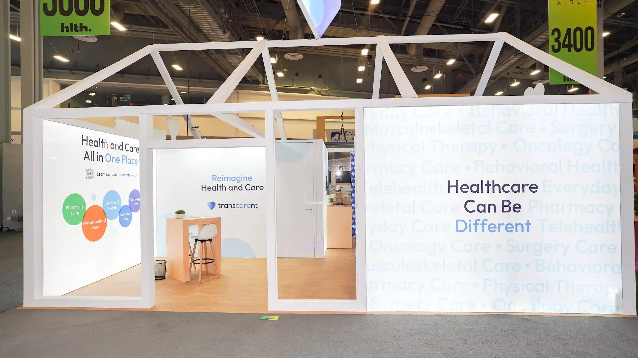 Exterior angled view of Transcarent house booth from different event showing left side face with One Place for Health and Care headline on watermark background, glowing lantern house effect against purple ambient convention lighting, roof truss shadow patterns on walls, heart sculpture at apex, warm wood interior floor visible through open frame, Health Gorilla and Bioformis neighbors visible