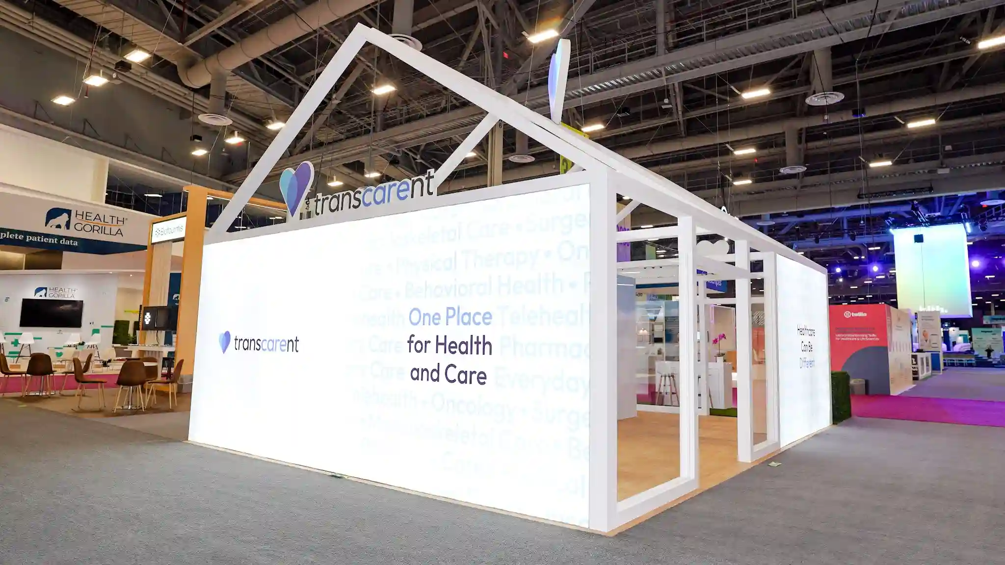 Front-left corner view of Transcarent house booth showing three face panels with different messages, two iridescent pearl-finish heart sculptures, Healthcare Can Be Different panel, branded door, Inovalon booth in background