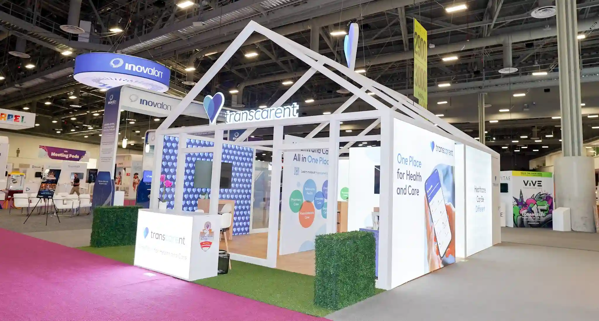 Wide-angle frontal-left view of Transcarent house booth at HLTH 2023 showing broadest view of complete booth with open house truss frame and gabled roof, heart-patterned interior walls visible inside, front left reception counter, multiple sequential graphic panels on right wall telling storyboard narrative, green grass flooring and boxwood hedge, pink HLTH aisle carpet, Inovalon and VITAE and VivE neighbors visible
