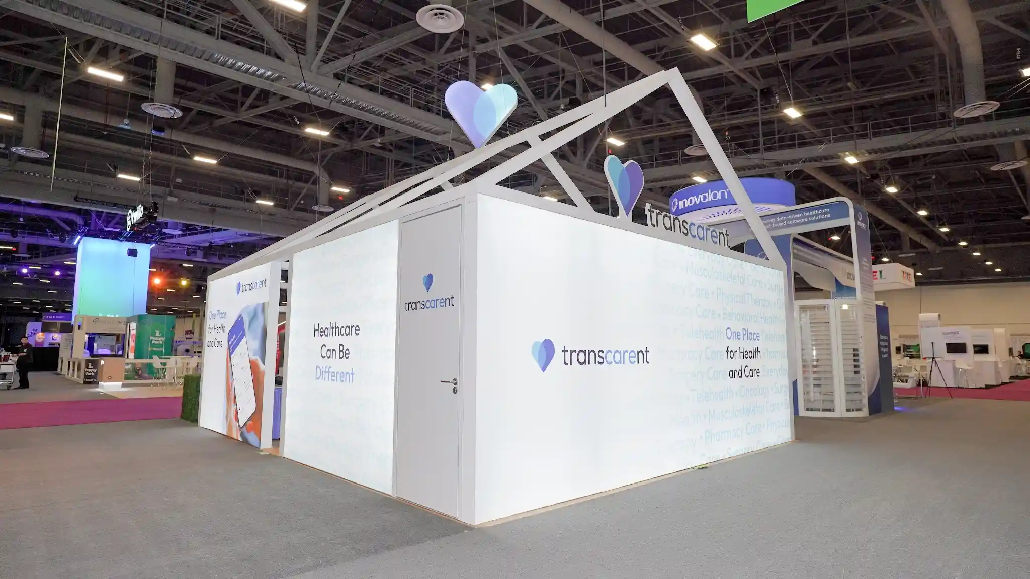 Wide-angle exterior view of Transcarent house booth from different angle showing full front and right side faces, two oversized 3D heart sculptures at roof apex and fascia, watermark typography wall, functional branded door, Inovalon neighbor