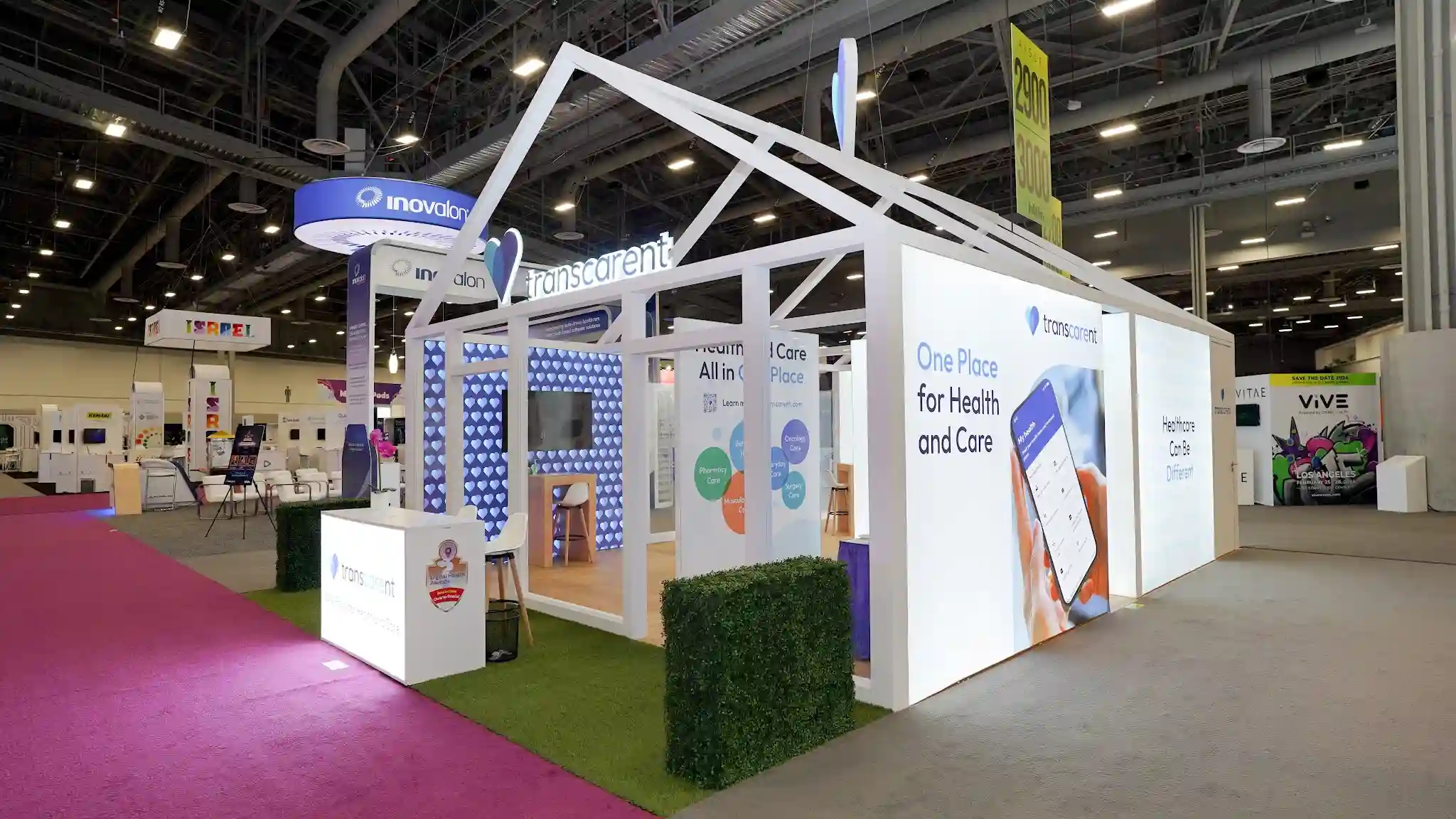 Wide establishing shot of the Transcarent 20x30 ft custom island exhibit at HLTH 2023 at the Las Vegas Convention Center West Hall showing the full house-shaped booth structure with dramatic white open-truss gabled roof frame forming a house silhouette above the show floor, large 3D heart sculpture in iridescent teal-blue tones suspended from the roof apex, brilliantly backlit white SEG fabric wall panels glowing like a lantern, Transcarent heart logo and wordmark on roofline fascia in blue, right side wall panel displaying One Place for Health and Care headline with smartphone app graphic showing My Health screen, Healthcare Can Be Different messaging, artificial green grass flooring creating front lawn perimeter, boxwood hedge planter cube at front exterior, pink aisle carpet of HLTH conference, front reception counter with Transcarent branding and award badge, neighboring Inovalon booth with blue circular hanging sign visible, HLTH aisle markers 2900 and 3000 visible in background