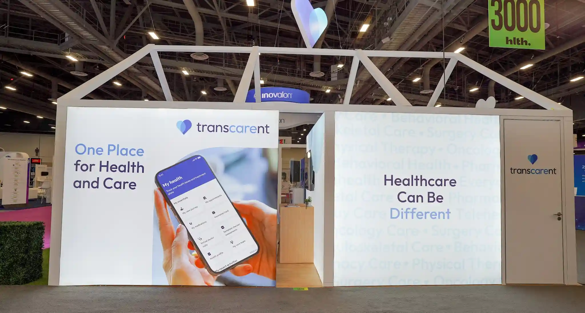 Wide-angle front-and-side perspective of the Transcarent 20x30 ft house-shaped island booth at HLTH 2023 showing three-quarters of the structure with open front face of house frame, full right side backlit wall, blue heart-patterned interior accent walls visible inside, Transcarent wordmark and heart logo on roofline fascia, right side wall with One Place for Health and Care headline and smartphone app graphic and Healthcare Can Be Different messaging, reception desk at lower-left front with Transcarent logo and Digital Health award medallion, warm wood-look interior flooring, green artificial grass perimeter, boxwood hedge planter, bar stools and counter-height tables inside, real white flush door set into right side wall, pink HLTH aisle carpet, Inovalon blue circular sign in background