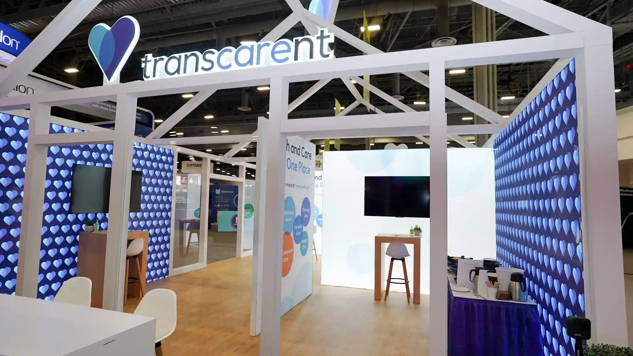 Straight-on front elevation of the Transcarent 20x30 ft house-shaped booth at HLTH 2023 showing full front facade with perfect house silhouette symmetry, white open-truss gabled roof frame with diagonal cross-bracing truss members forming architectural roof skeleton with triangular peak, small 3D teal-blue heart sculpture visible suspended from roof apex, left wall panel with Transcarent heart logo and wordmark and One Place for Health and Care headline and large smartphone graphic showing Transcarent mobile app My Health screen with menu items My essentials My appointments My care journey Insurance Card My medications, right wall panel with Healthcare Can Be Different messaging with Different in lighter blue and subtle watermark health service terms, far right small panel with Transcarent logo above flush lever-handle door, backlit panels glowing luminously, HLTH 3000 aisle sign in background, Inovalon booth visible