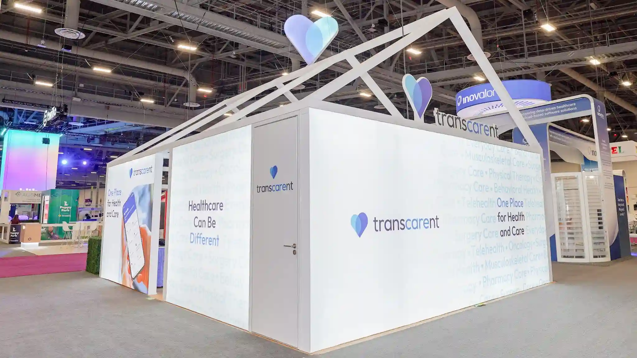 Interior corridor view of Transcarent booth showing private meeting room door with Reimagine Health and Care messaging, blue bubble watermark pattern on wall, warm wood flooring