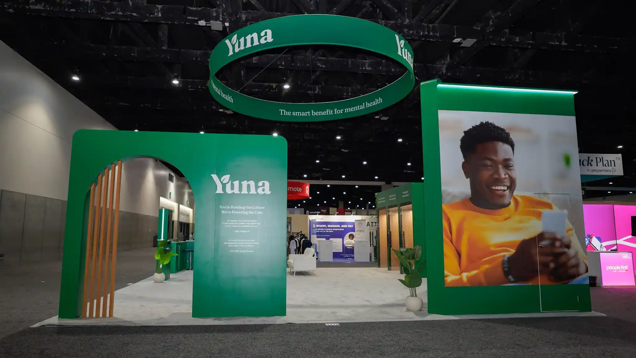 Front entrance of Yuna booth with large green archway featuring natural wood slats, You're Building the Culture We're Powering the Care messaging, circular LED halo hanging sign, lifestyle graphic pylon, and potted plants at SHRM 2025