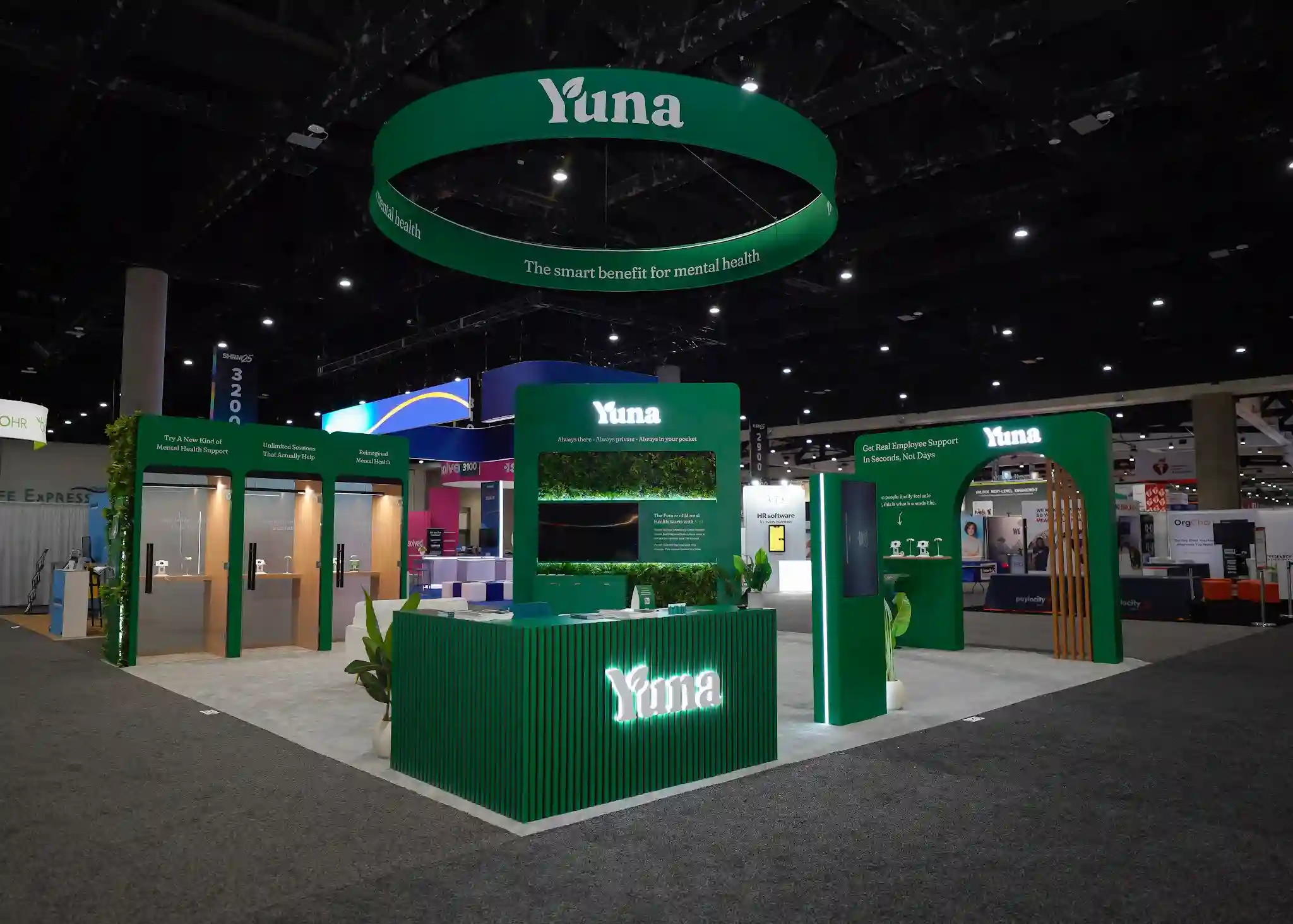 Yuna reception desk with vertically slatted green texture and glowing Yuna logo, living moss and greenery wall, The Future of Mental Health Support information panel, demo screen, and green stool