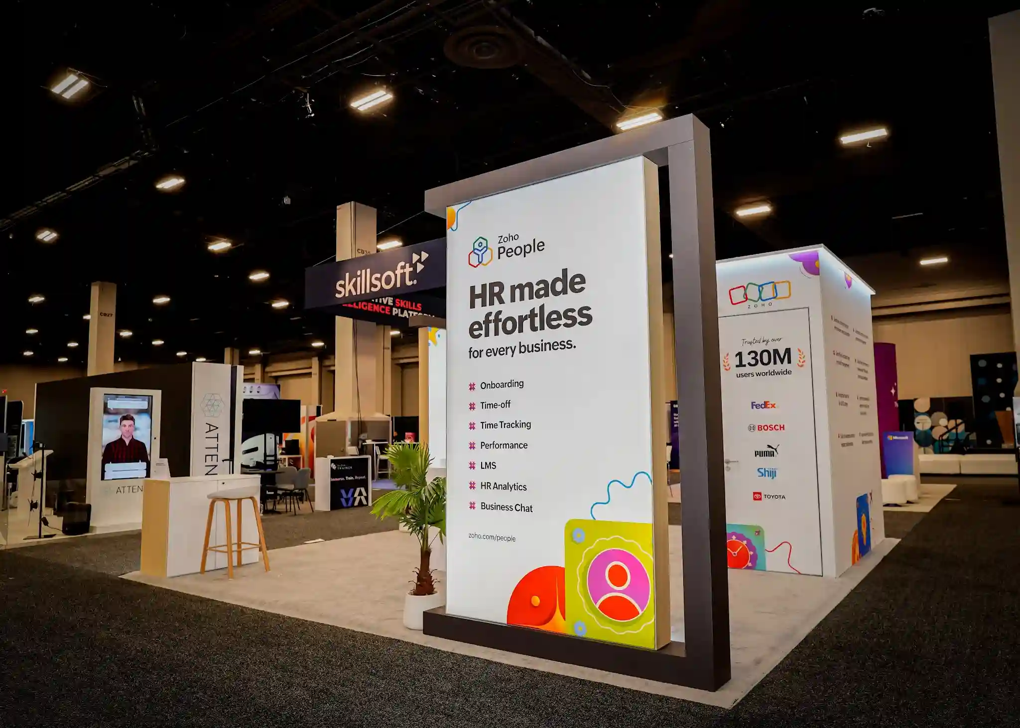 Close-up of the Zoho People branded lightbox display panel showing HR made effortless for every business headline with feature icons for Onboarding Time-off Time Tracking Performance LMS HR Analytics and Business Chat, colorful illustrated icons at the bottom, dark charcoal frame structure, and tropical palm plant in white planter at base